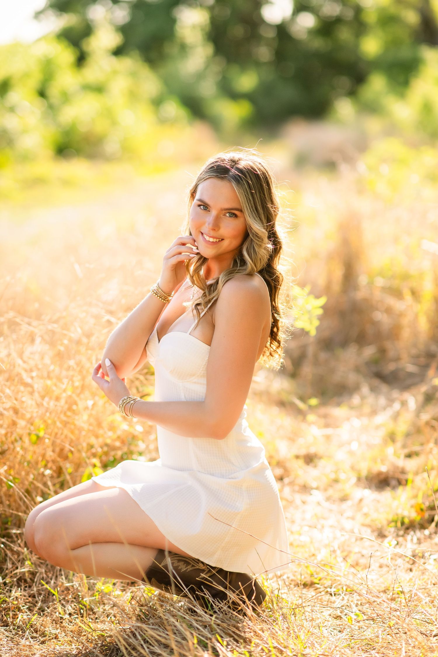A woman poses gracefully in a white sundress amidst tall golden grass in warm sunlight.
