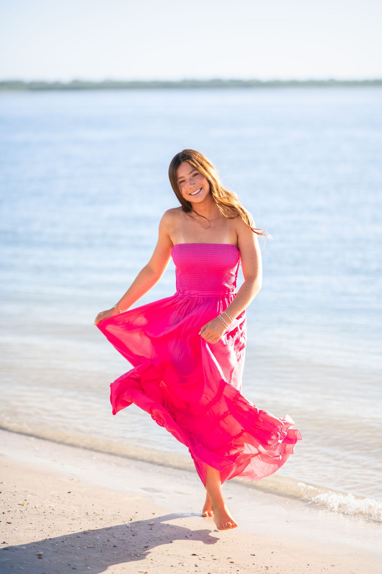 Woman in flowing bright pink dress twirls joyfully on sunny beach against blue ocean backdrop.