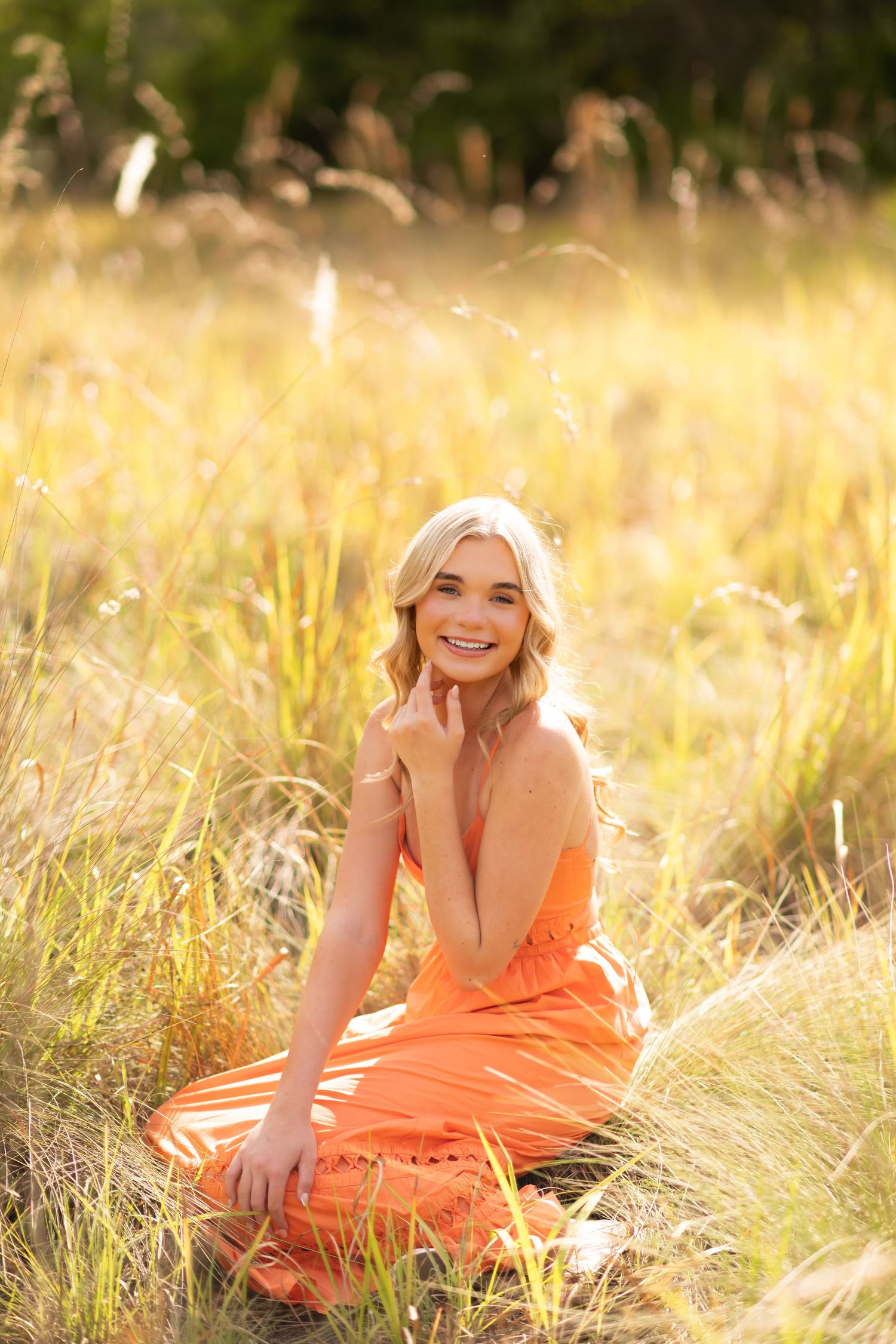 Young blonde woman in orange dress sitting in golden field, smiling in warm sunlight.