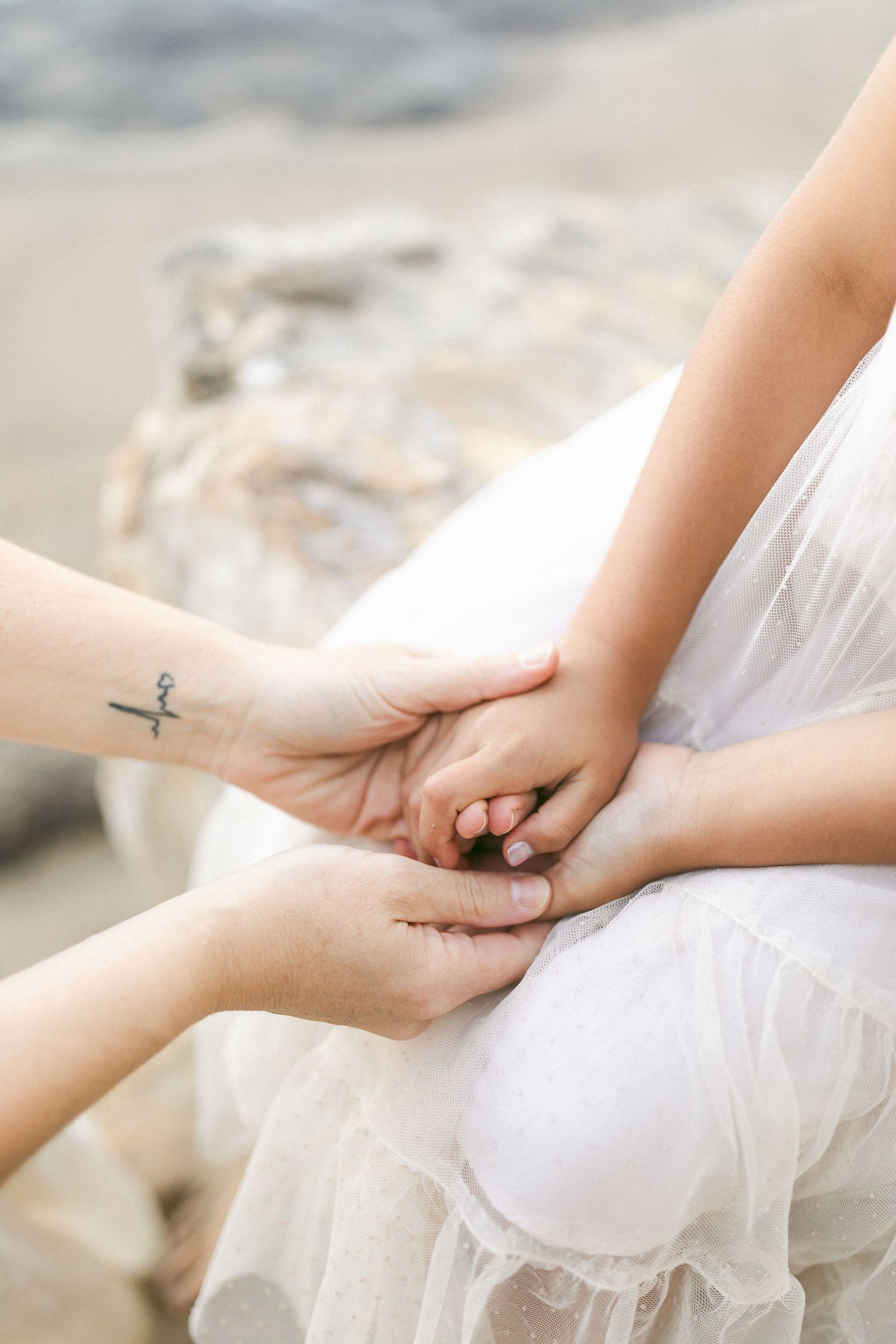 Two hands clasp together tenderly over a flowing white dress, with one person showing a small tattoo on their wrist captured by Chelsee Raw.