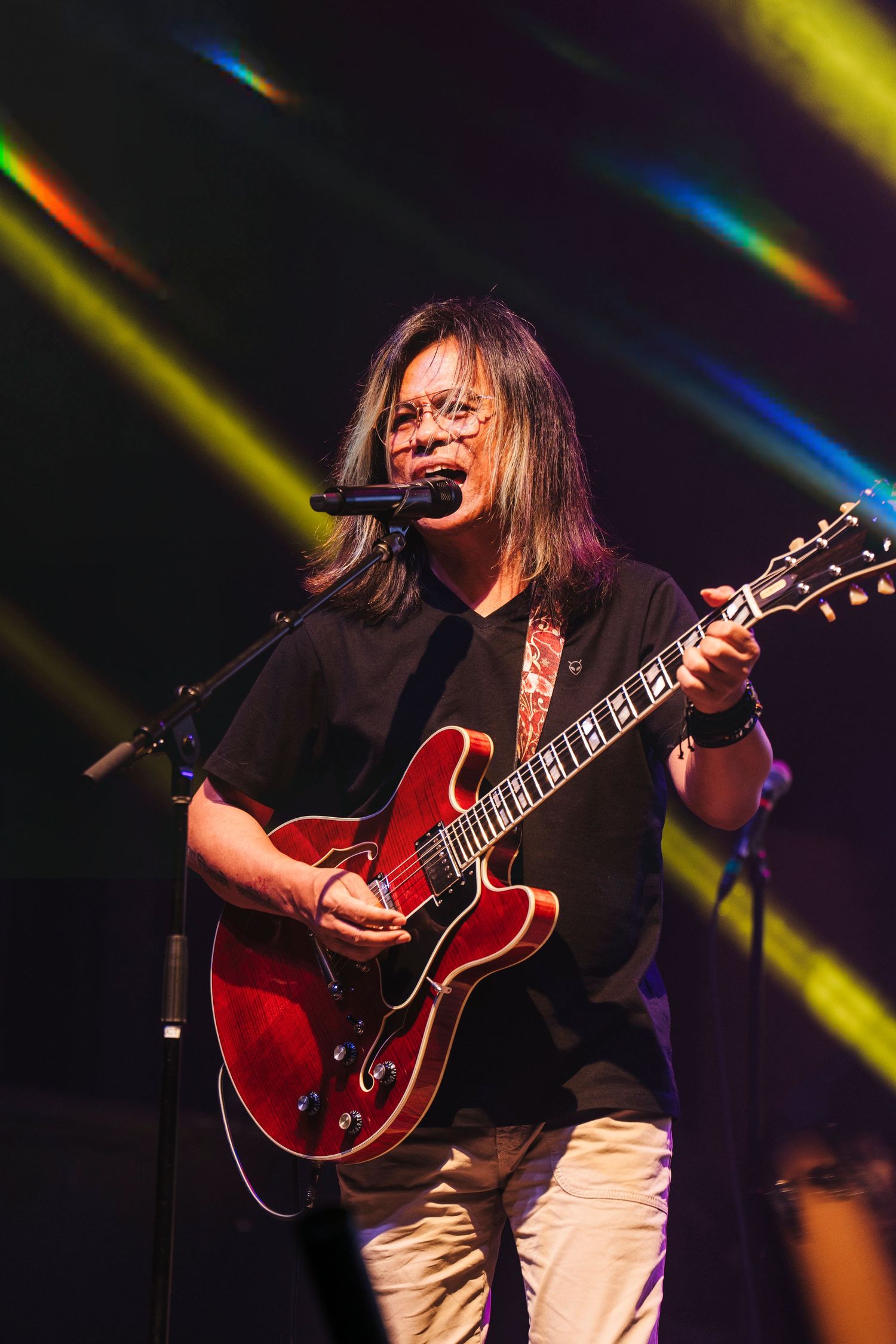 Musician in black performing on stage with red guitar under colorful spotlights during live concert.