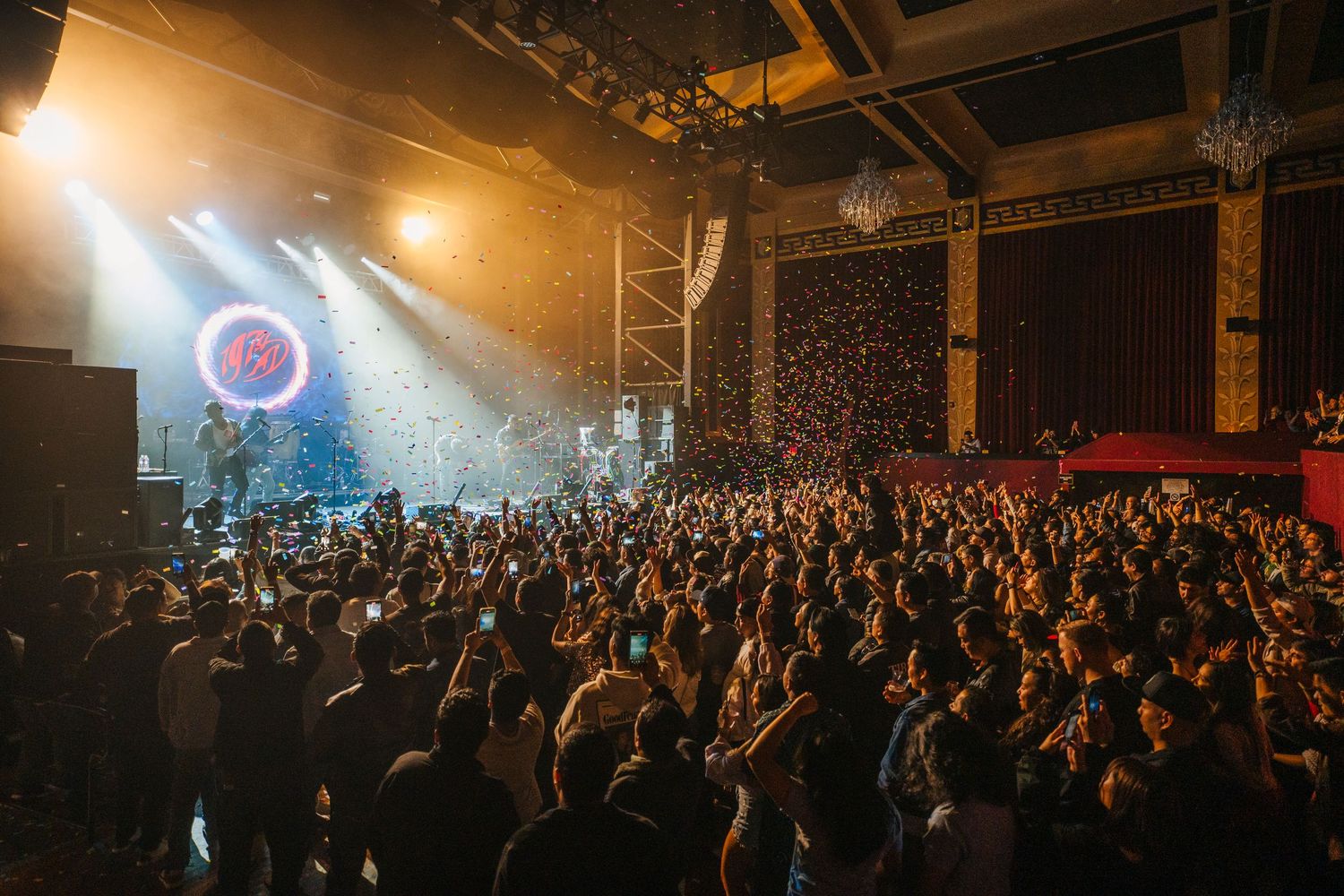 Large concert crowd inside a dark venue with dramatic spotlights and a glowing circular stage element illuminating the atmosphere.