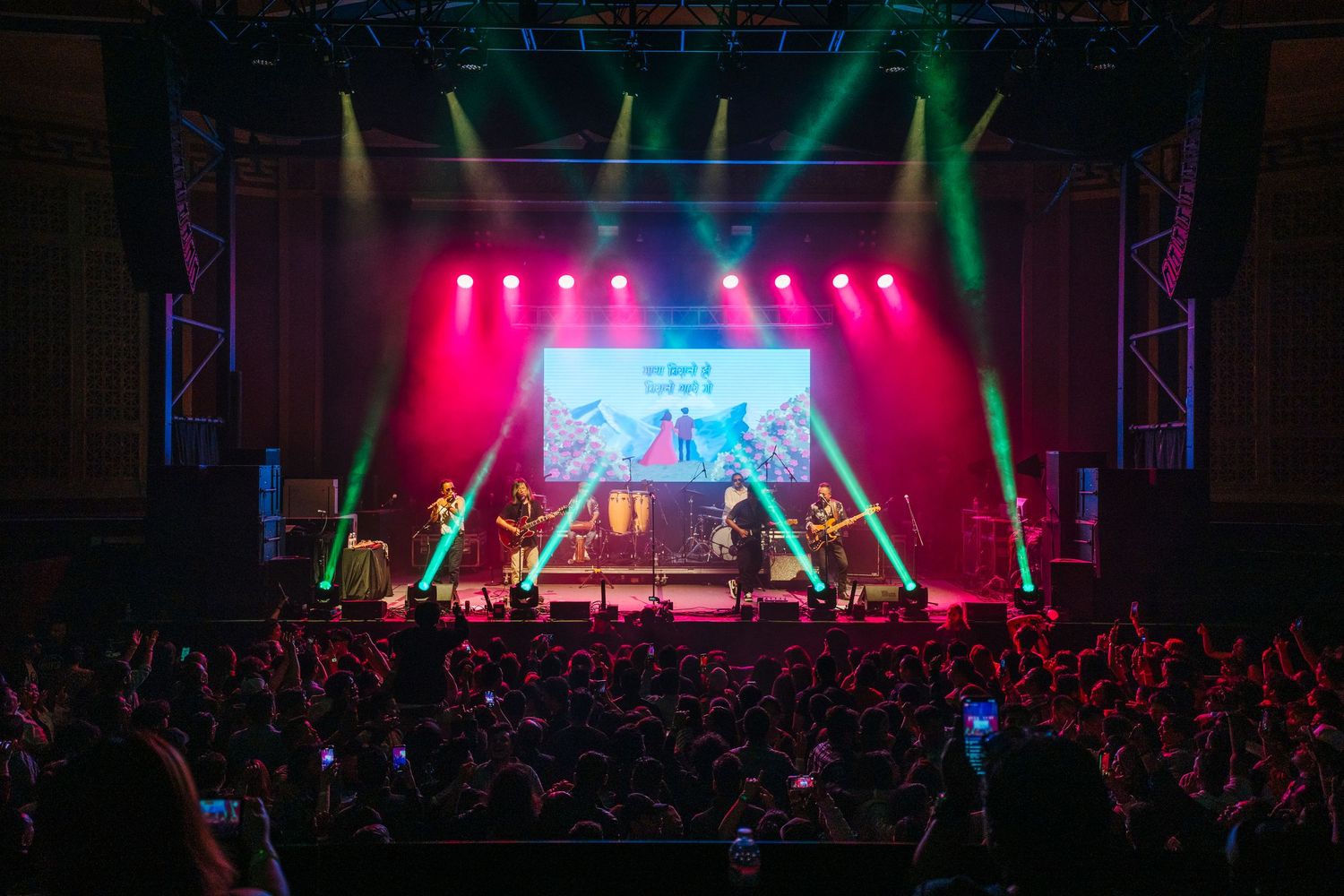 Concert stage illuminated with pink and green lights as performers entertain an energetic crowd at a live music show.