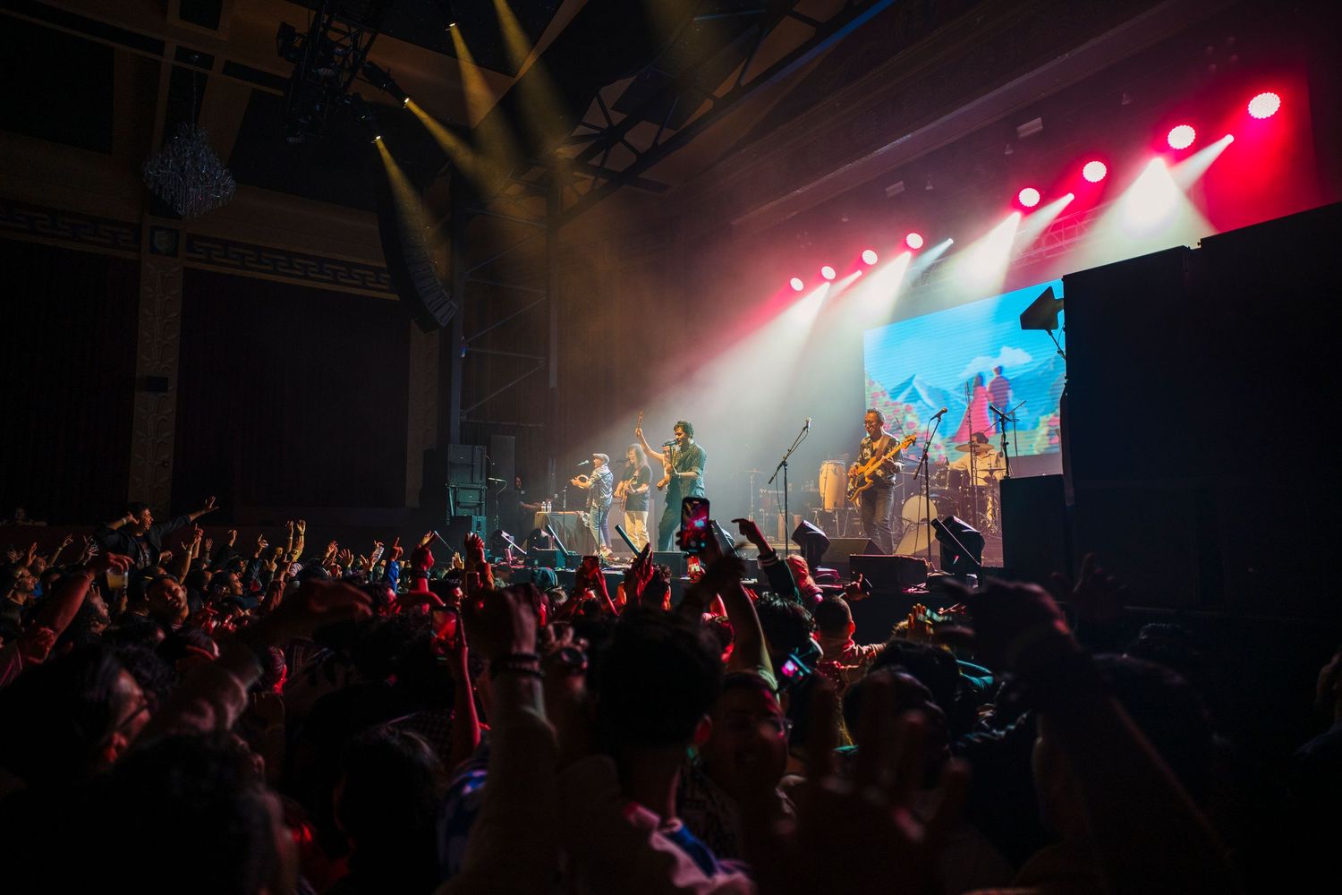 Concert crowd silhouettes raise hands in the air under colorful stage lighting during a live music performance.