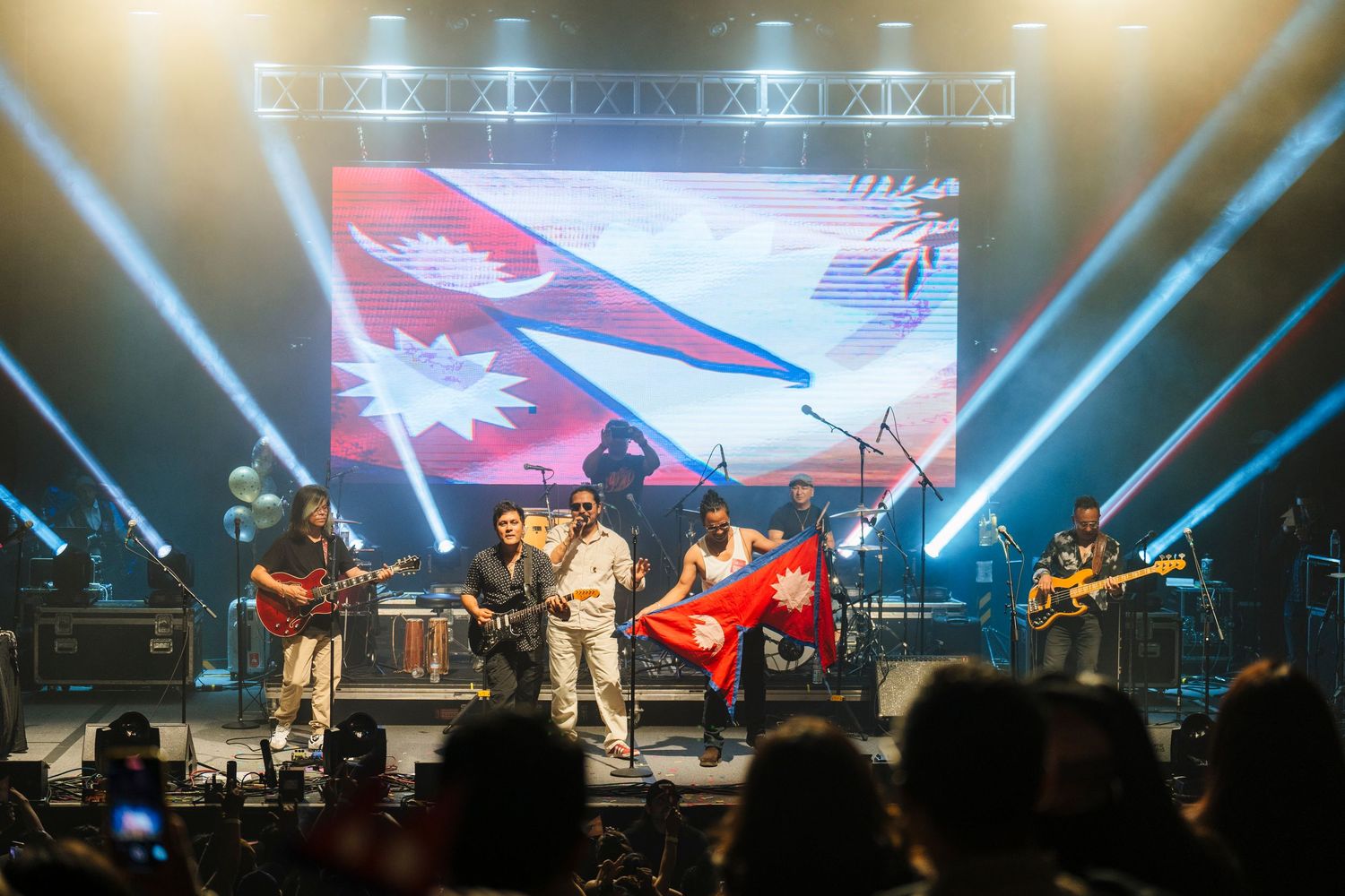 Concert stage lit with dramatic blue lights and video screens showing a red design as musicians perform for a crowd.