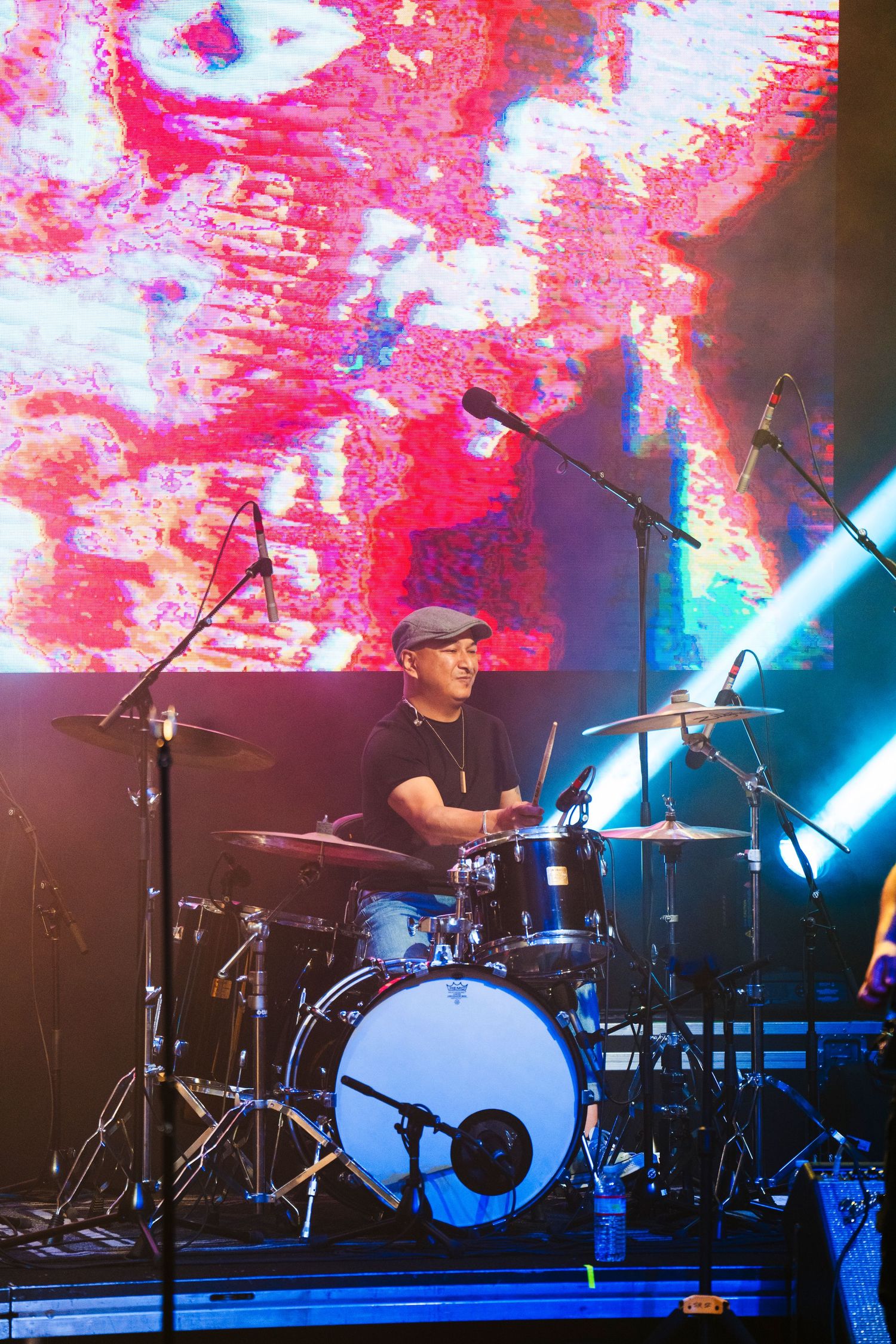 A drummer performs on stage with vibrant red and blue lighting and a large visual display behind the drum kit.