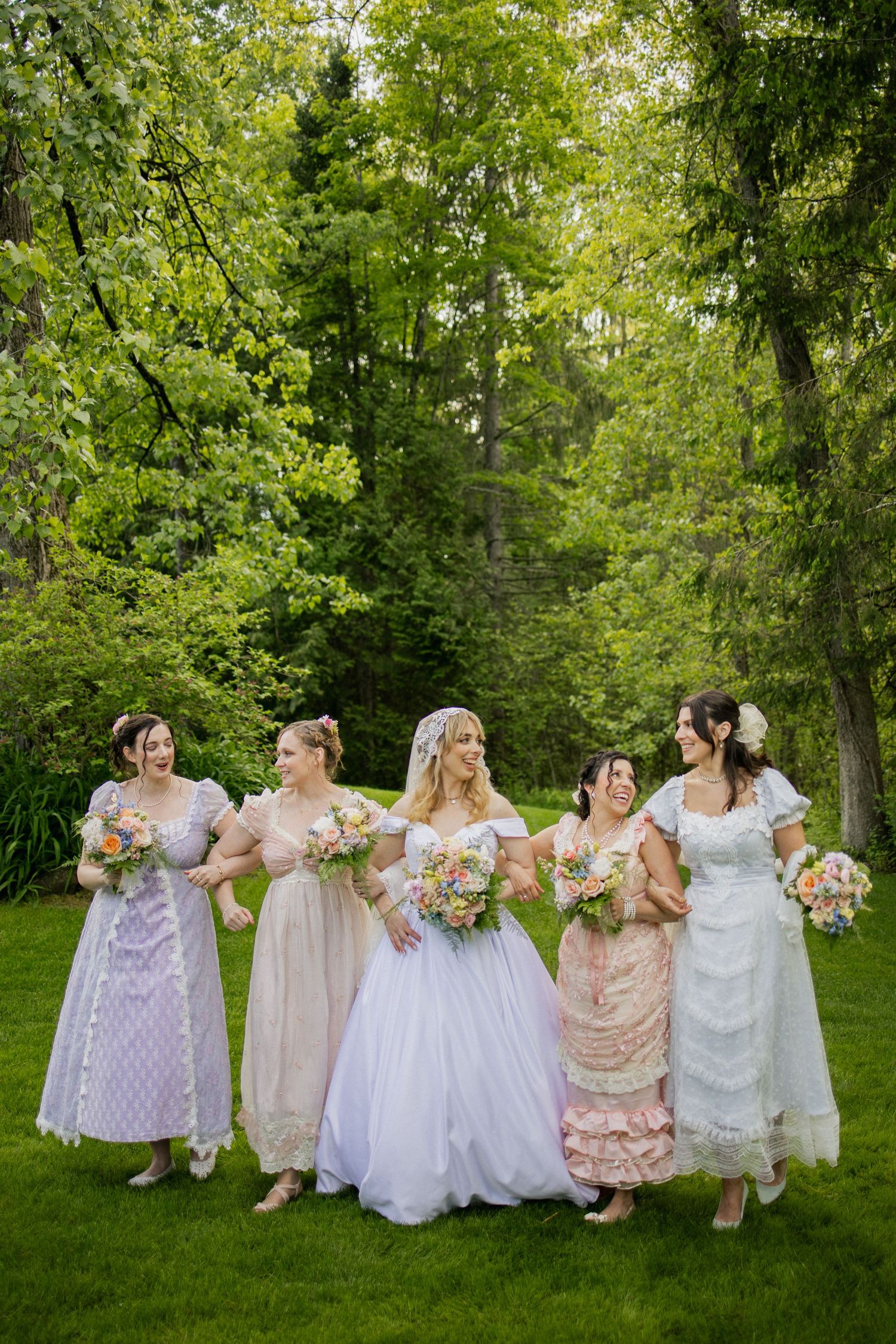 A group in pastel and white dresses sit together on grass during an outdoor wedding photography session.