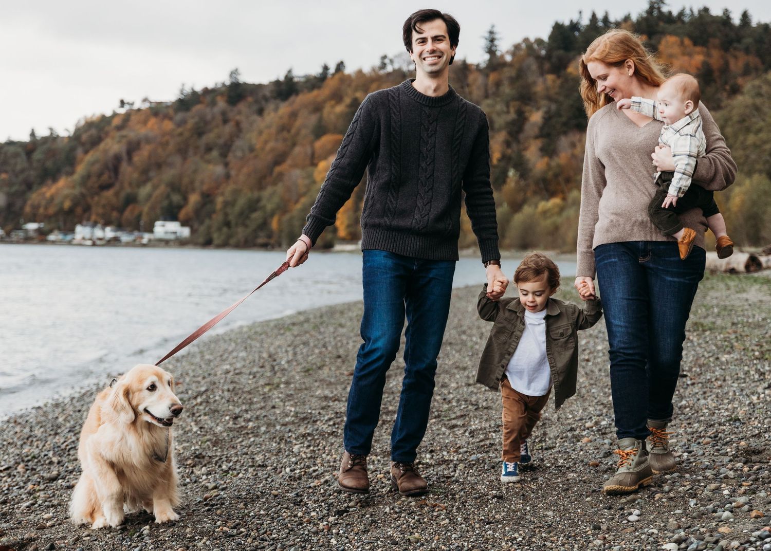 A couple walks with their playful golden retriever dog along a rocky waterfront beach on an autumn afternoon.