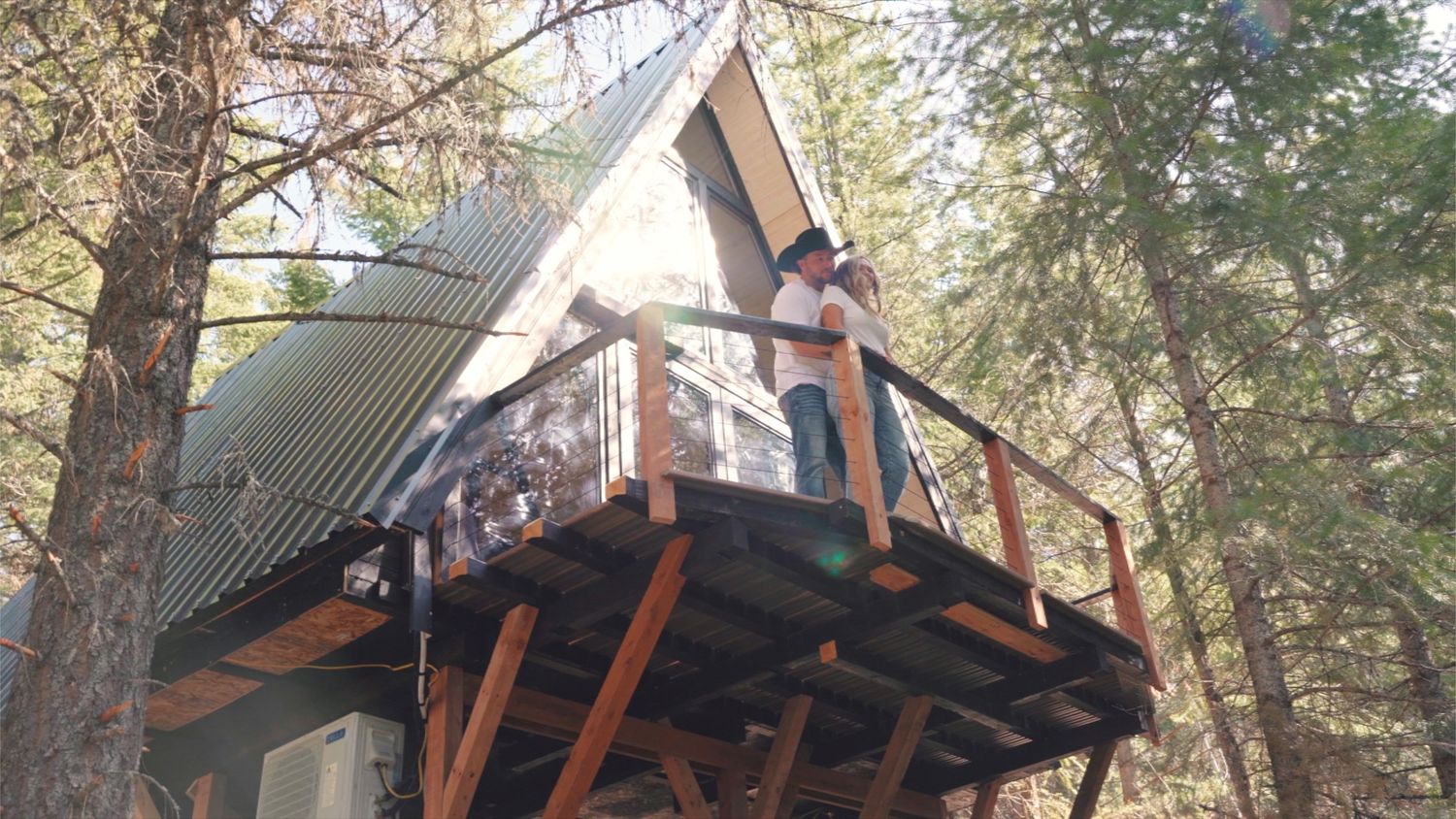 A rustic A-frame treehouse with wooden deck and railings nestled among tall pine trees in a forest setting.