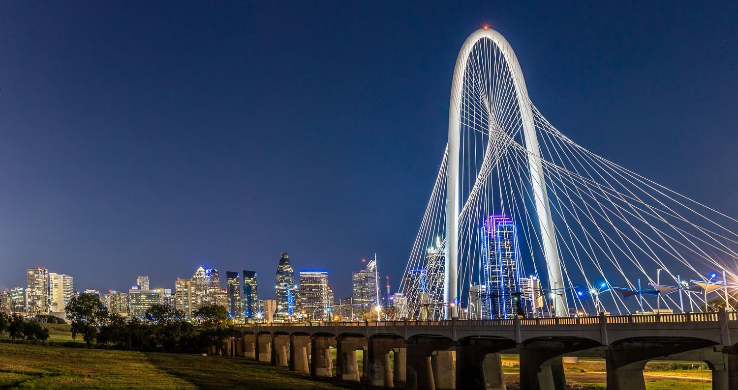 The illuminated Margaret Hunt Hill Bridge spans the Trinity River against the Dallas skyline at night.