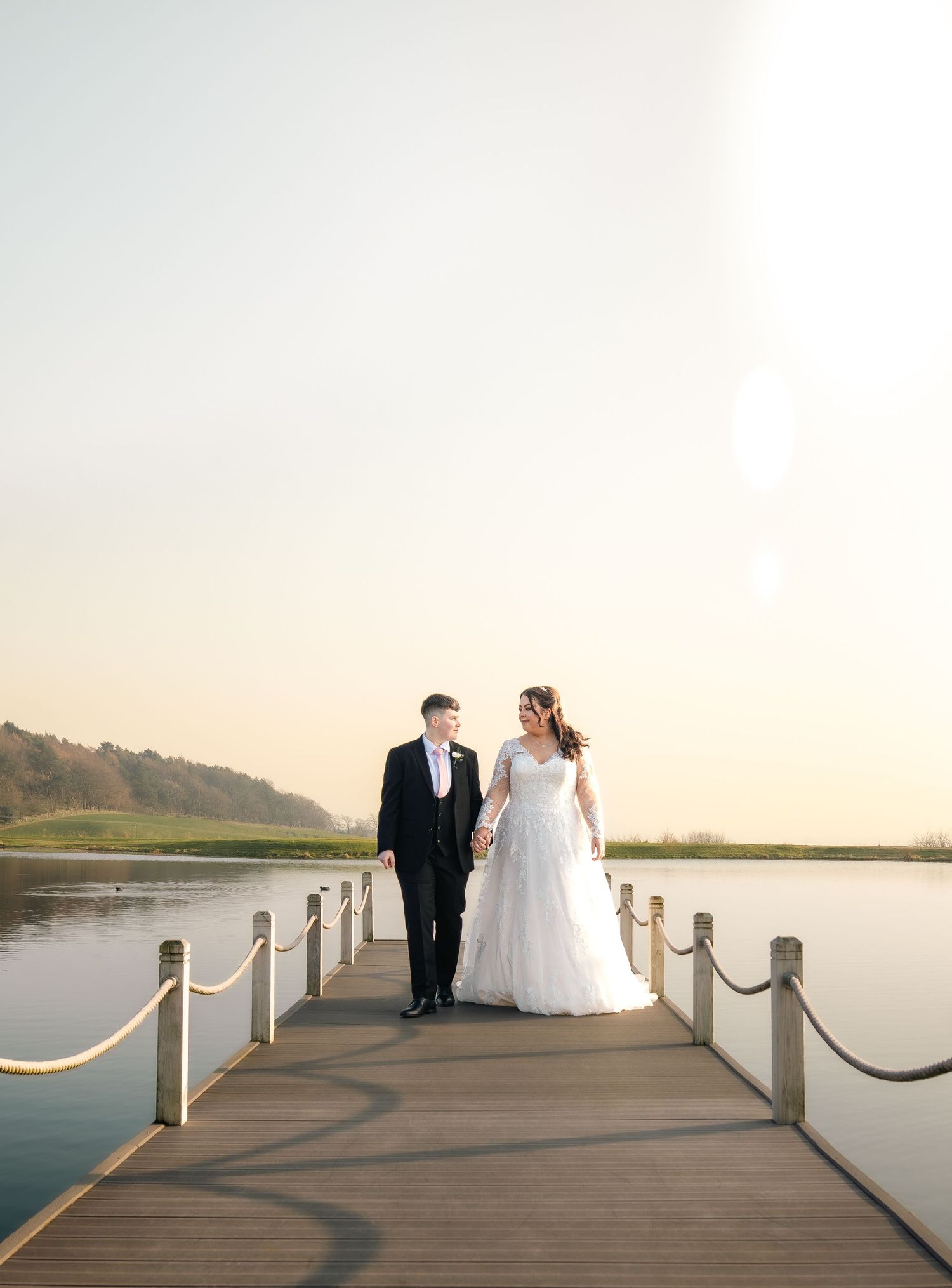 A couple in formal wedding attire stands on a wooden dock overlooking a serene lake at sunset.