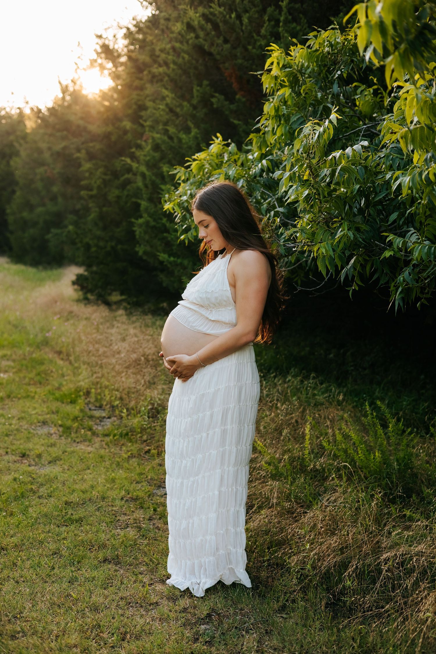 Expectant mother in white dress stands in garden during golden hour photography session.