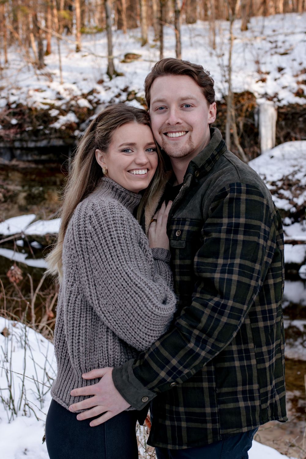 A couple in winter attire embraces in a snowy forest setting.