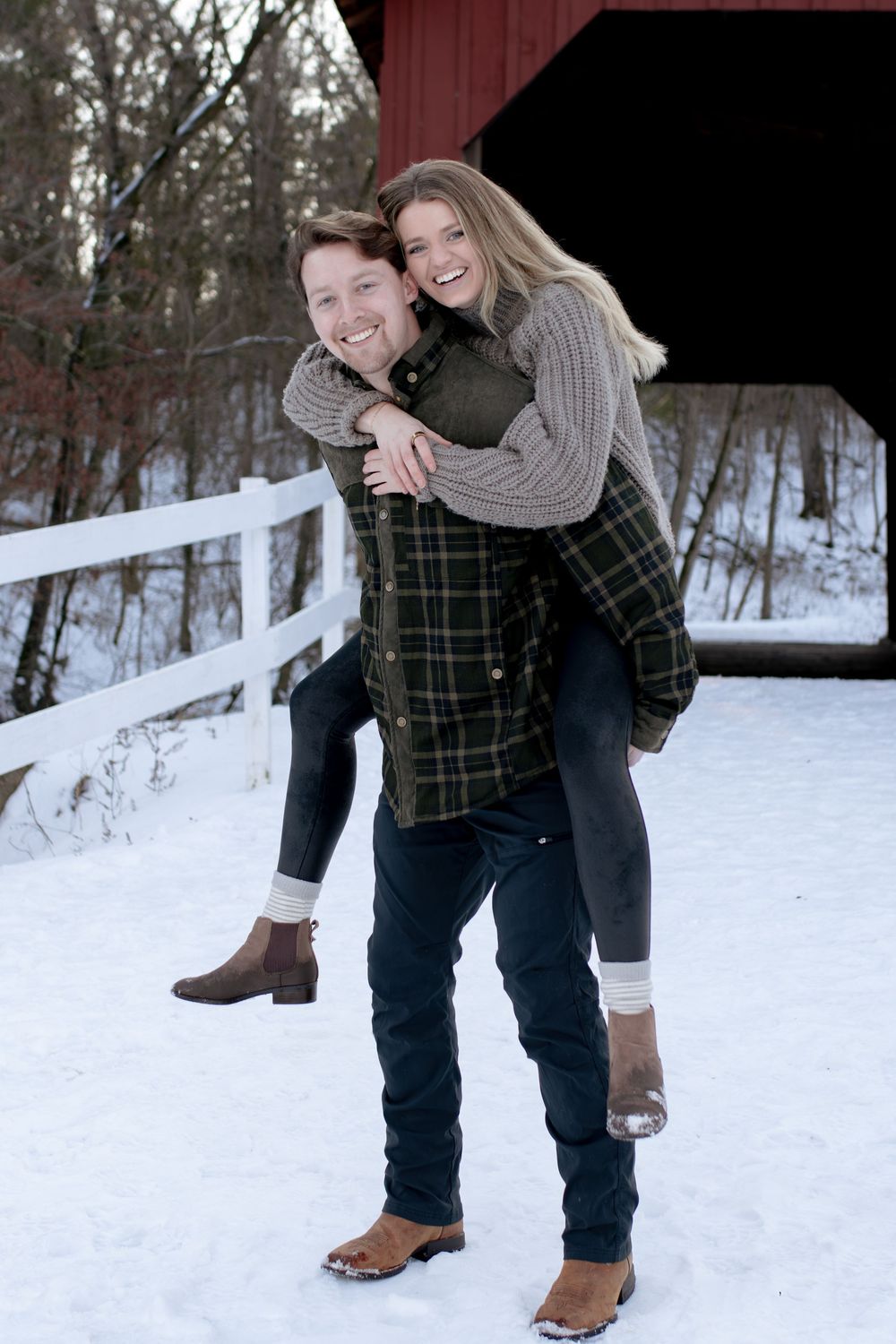 A playful winter photoshoot featuring a piggyback ride in front of a red barn with snow-covered ground.