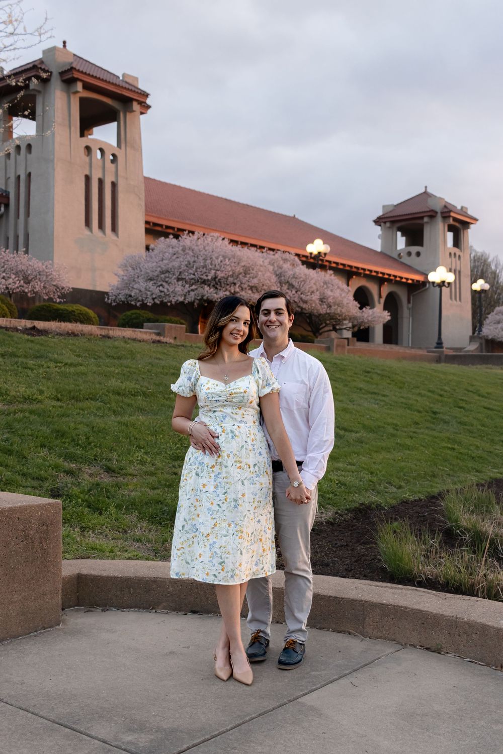A couple poses together outside a Spanish-style building with cherry blossoms in bloom.