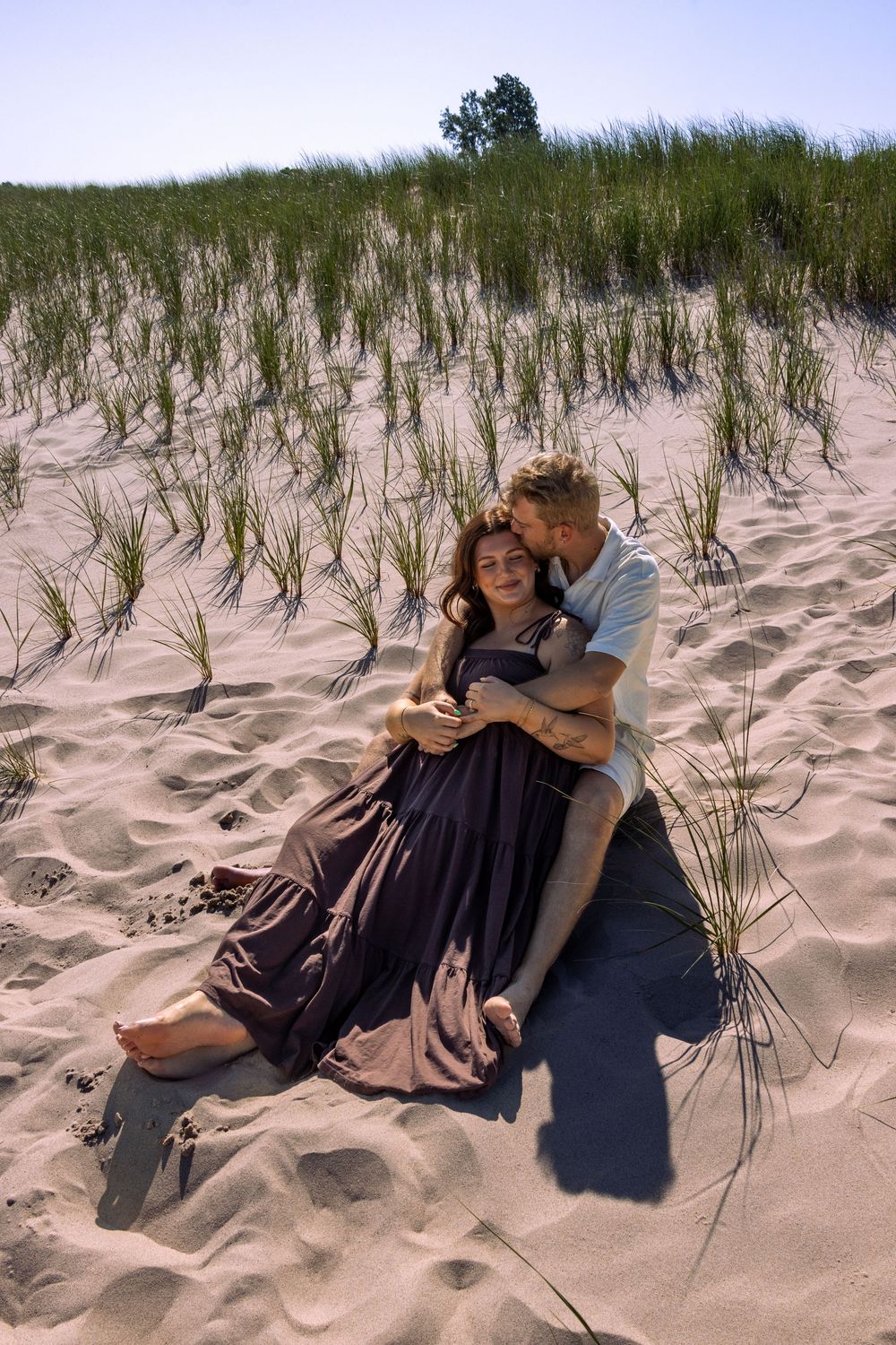 A couple embraces on sandy dunes with beach grass swaying in the background.