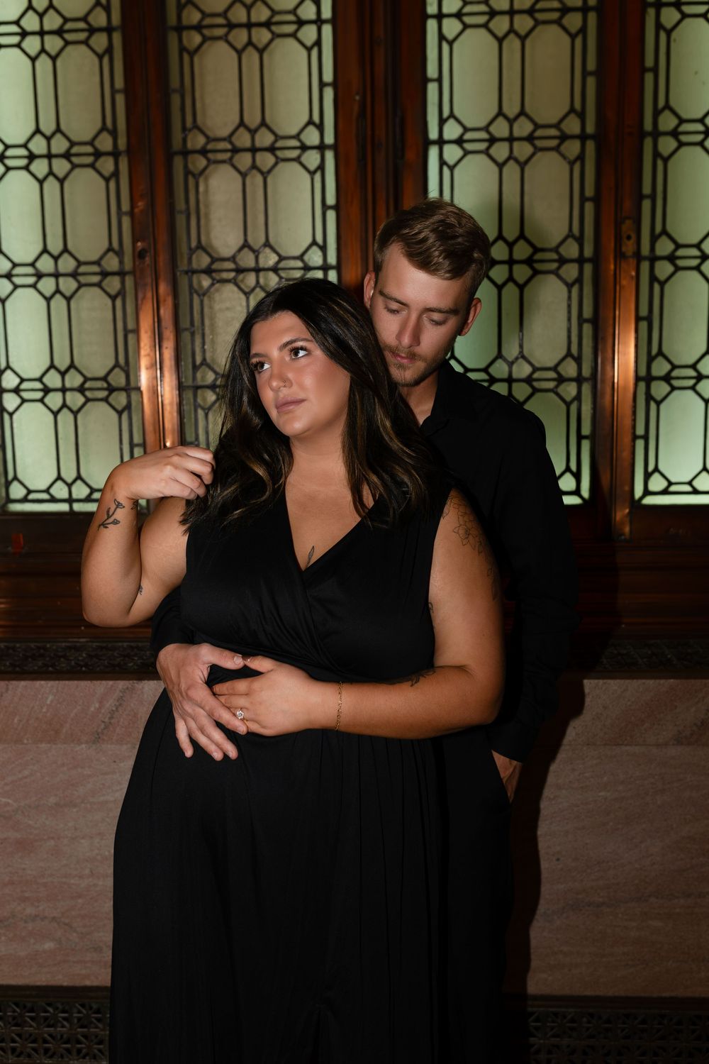 A couple in formal black attire poses together on ornate steps with decorative windows.
