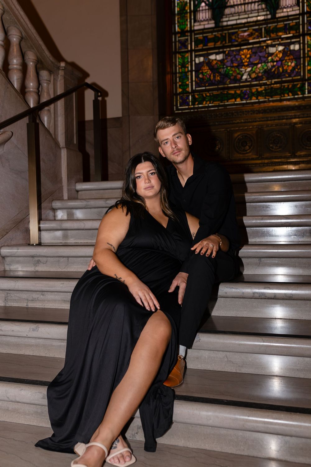A couple sits together on marble steps with stained glass windows in the background.