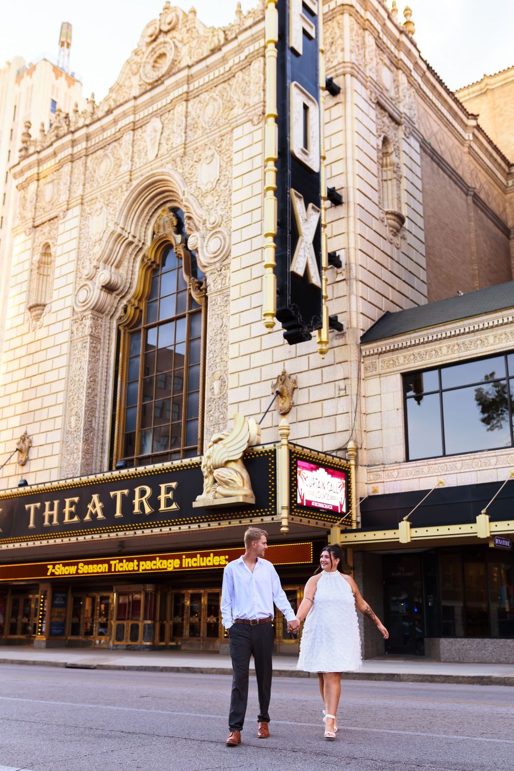 A couple walks hand in hand outside a classic theatre with ornate architecture.