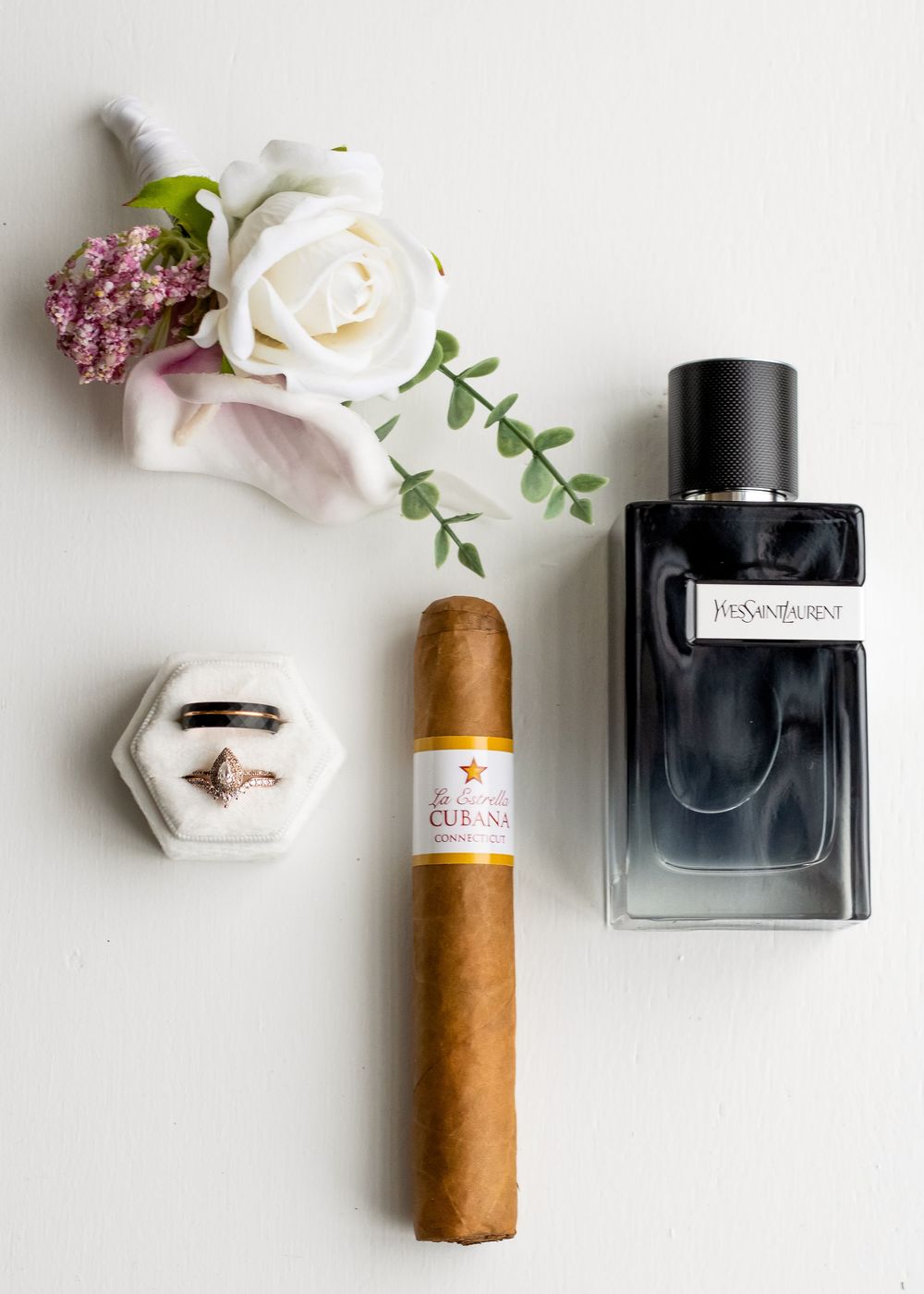 Wedding details flatlay with white rose, cologne bottle, and cigar on white surface.