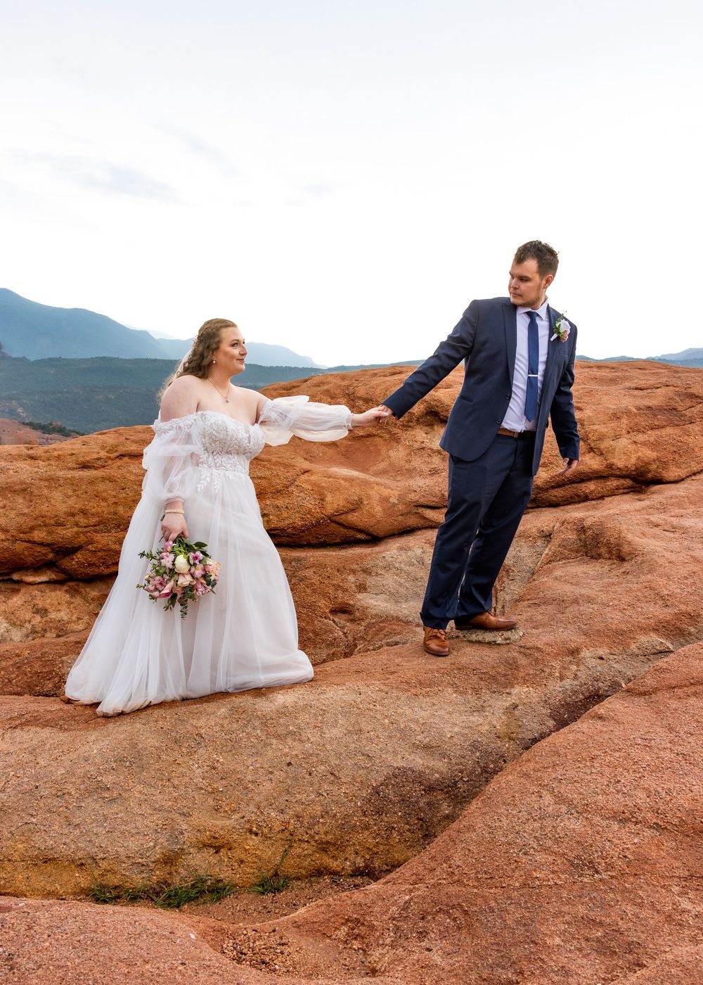 A playful wedding couple holds hands on red rocks with mountains in the background.