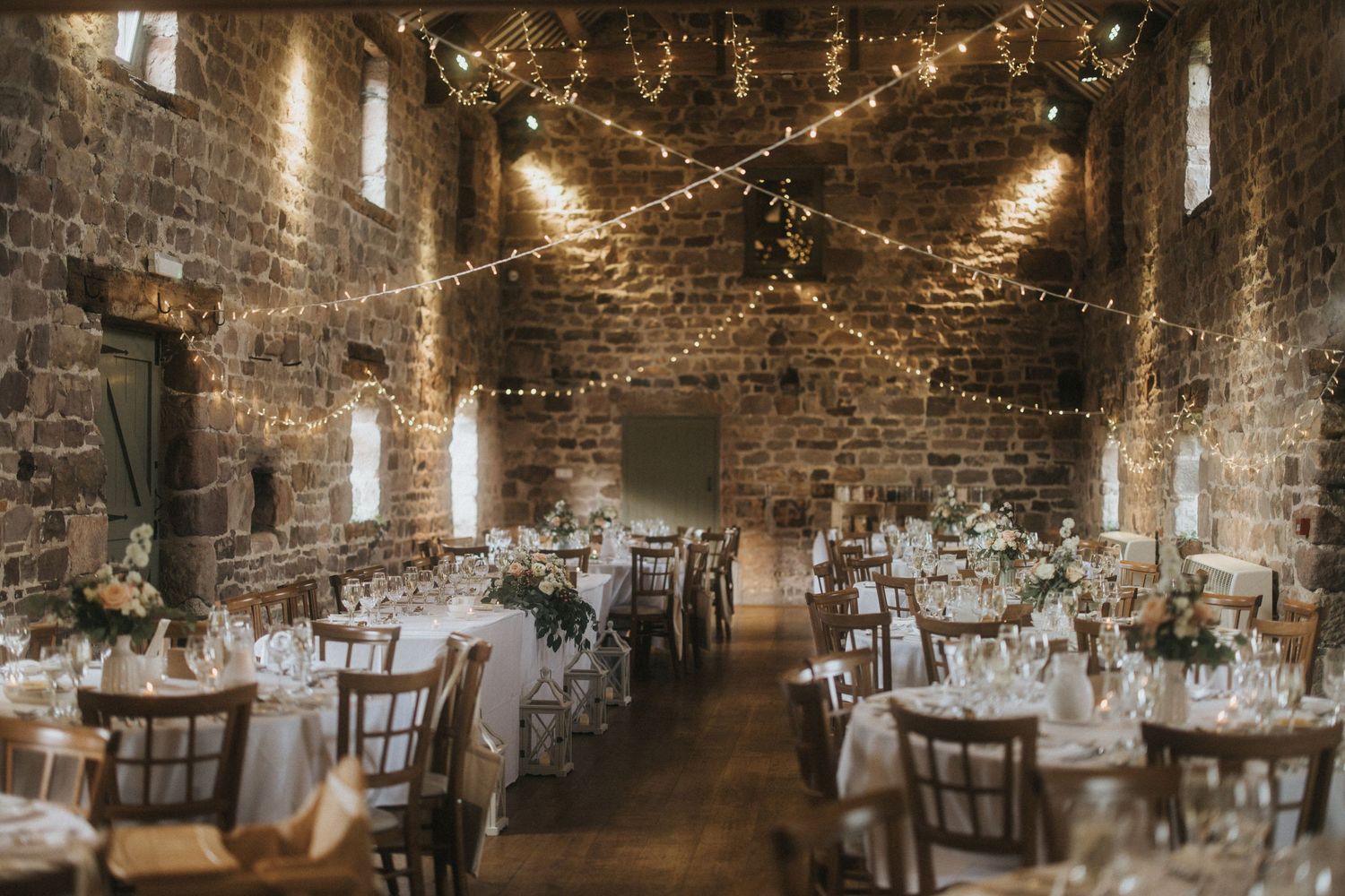 Rustic wedding reception venue with exposed brick walls, string lights, and elegant table settings in a historic barn space.