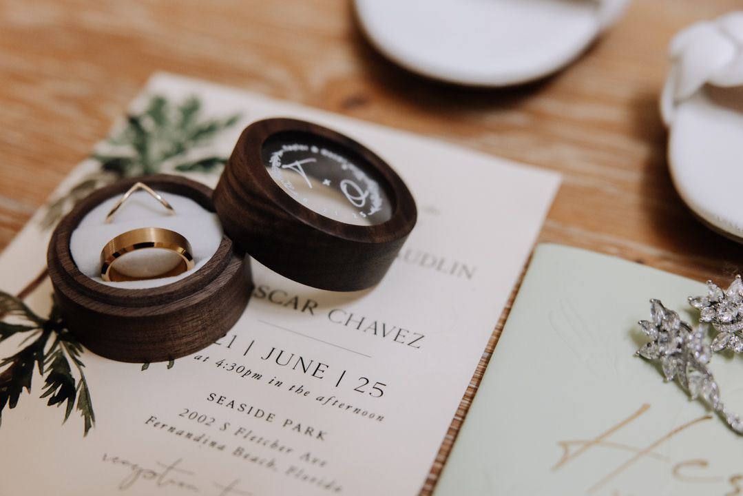 Wedding rings and invitation with greenery details photographed on a wooden surface in warm natural lighting.