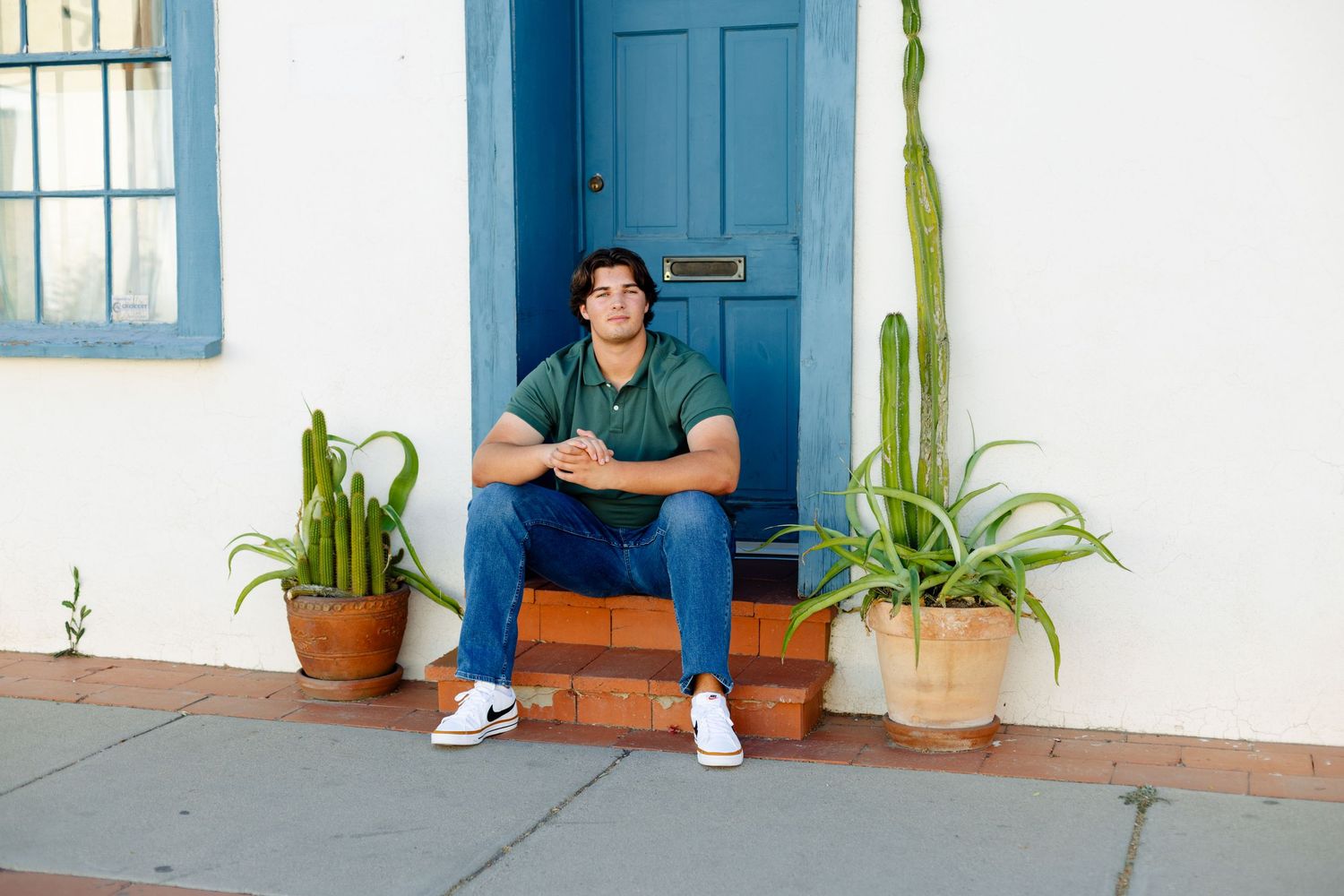 Person in casual attire sits on steps by blue door with potted cacti.