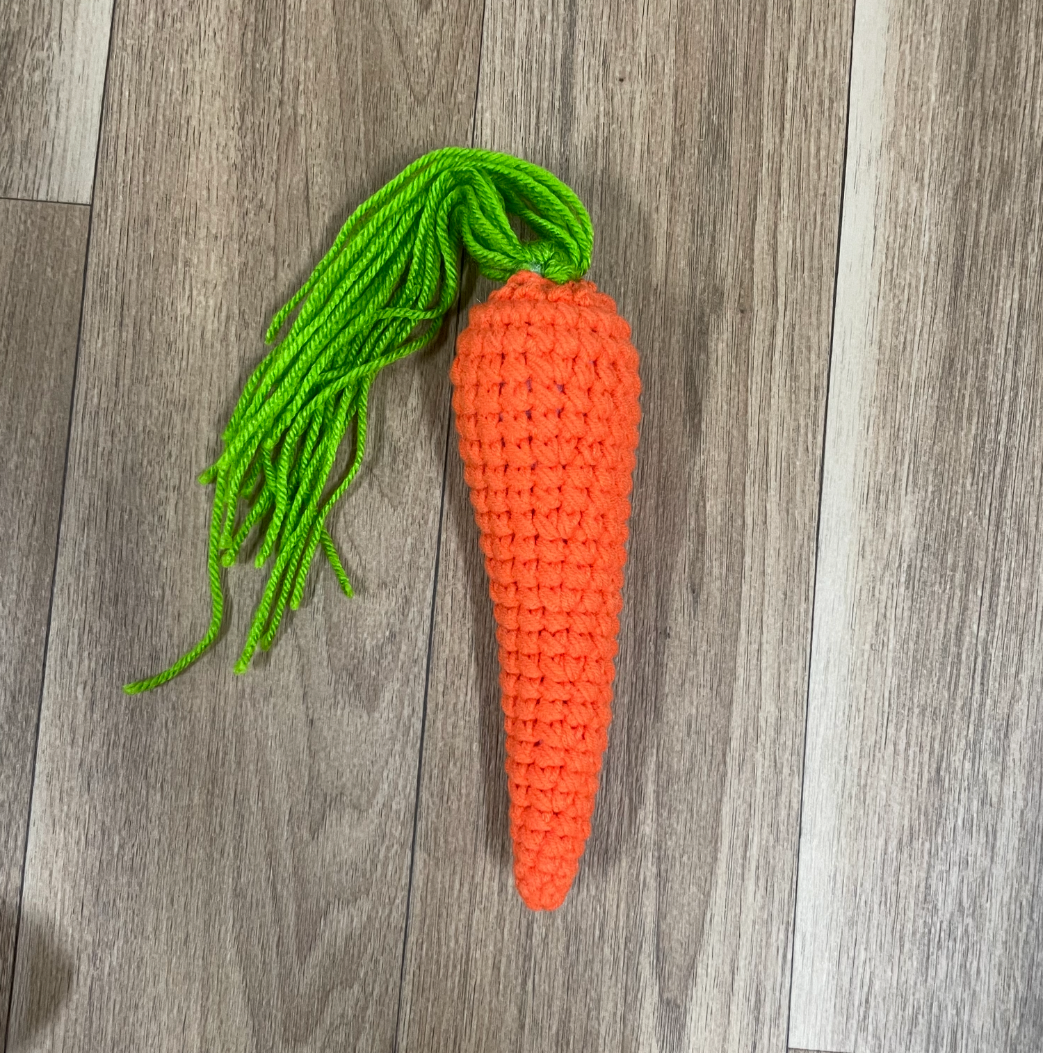 Decorative orange crochet carrots with green yarn tops hanging against a wooden floor background.