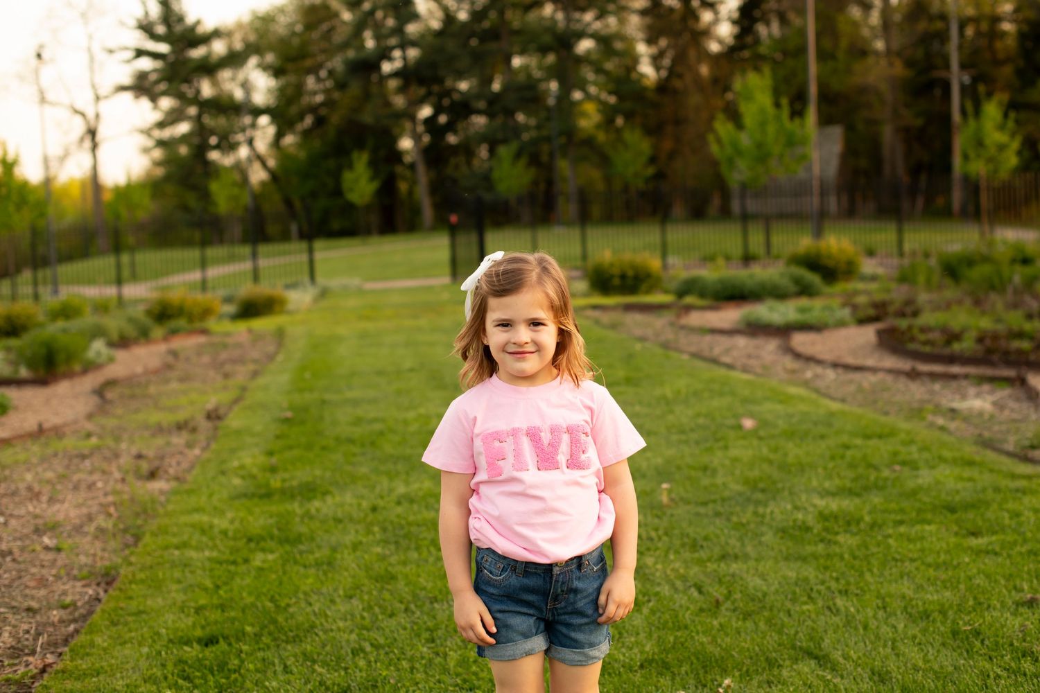 Young child wearing a pink t-shirt and denim shorts stands on a garden path with evergreen trees in the background.