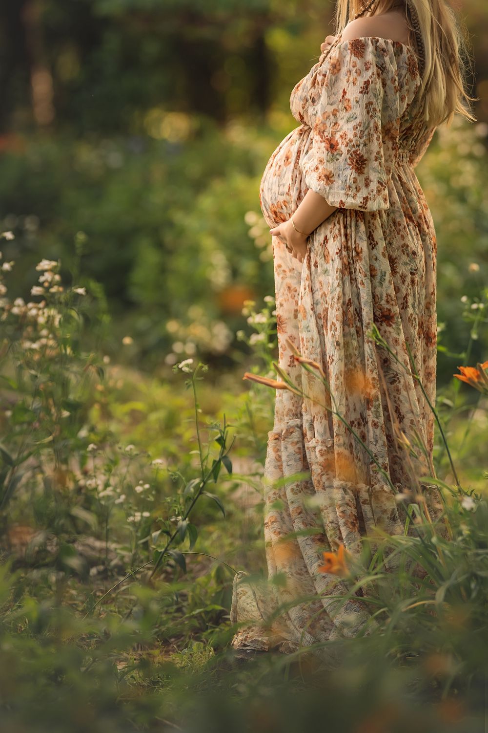 Expectant mother in floral dress stands among wildflowers in a sunlit garden during golden hour for Lookout Mountain Maternity photos.