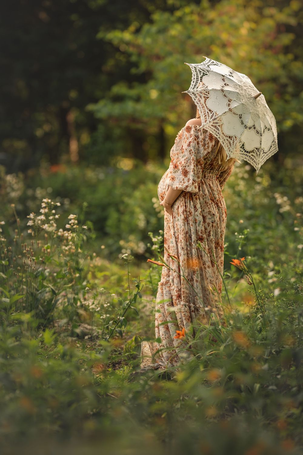 Woman in a floral dress and cintage parasol stands among wildflowers in a dreamy woodland garden setting for maternity photos in Chattanooga