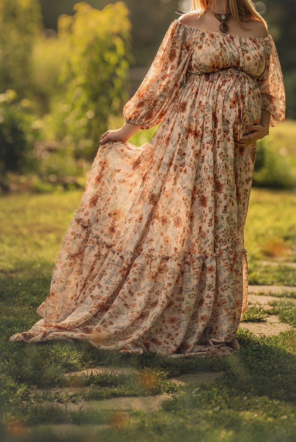 A flowing floral maternity dress billows gracefully in the sunlight while a pregnant woman stands on path in a garden setting.