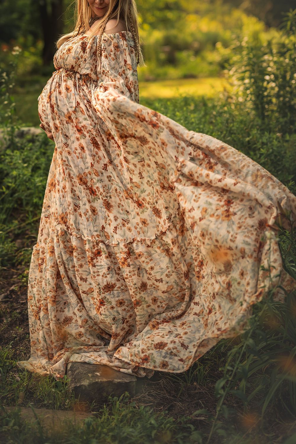 A flowy floral maternity dress billows in the wind against a lush green garden background during golden hour.