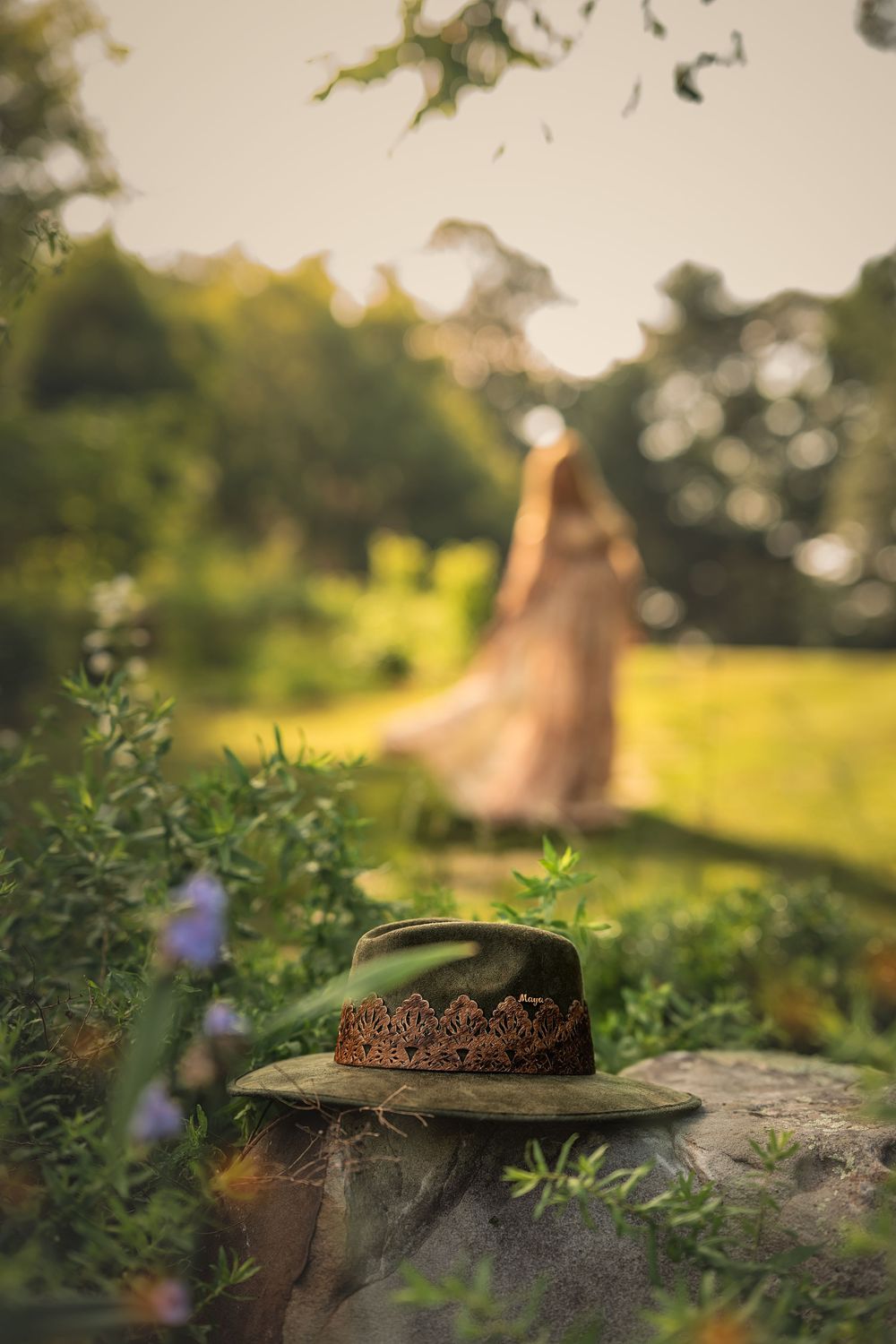 A green hat sits on a rock in a garden during golden hour lighting with a pregnant woman dancing in the bokeh behind.