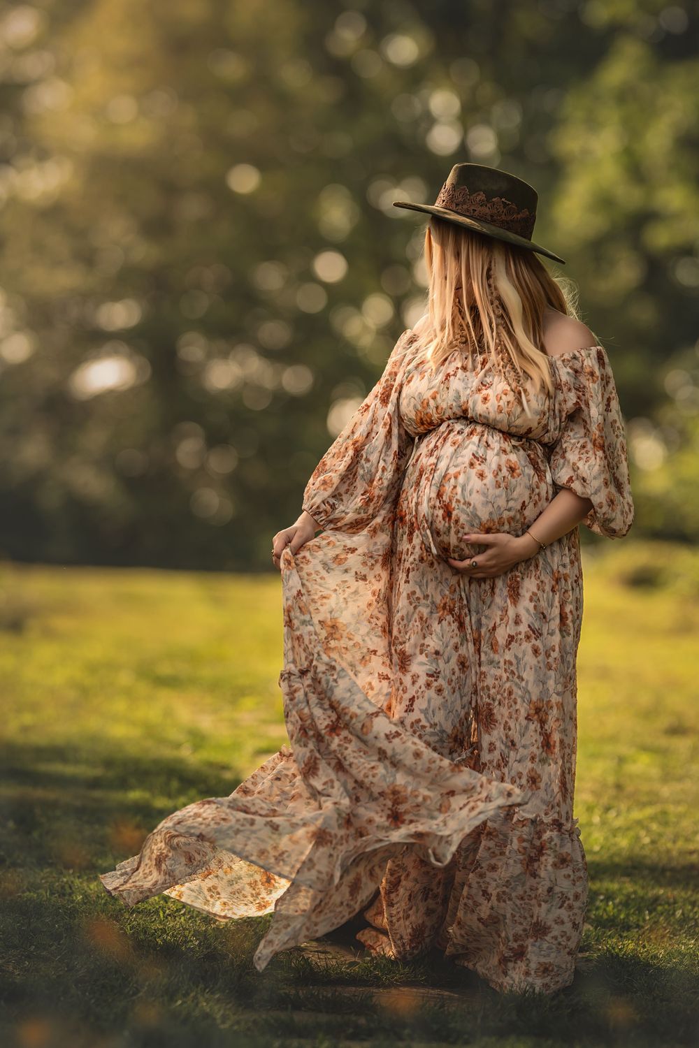 Pregnant woman in flowing floral dress and green hat stands in sunlit meadow with a golden bokeh background.