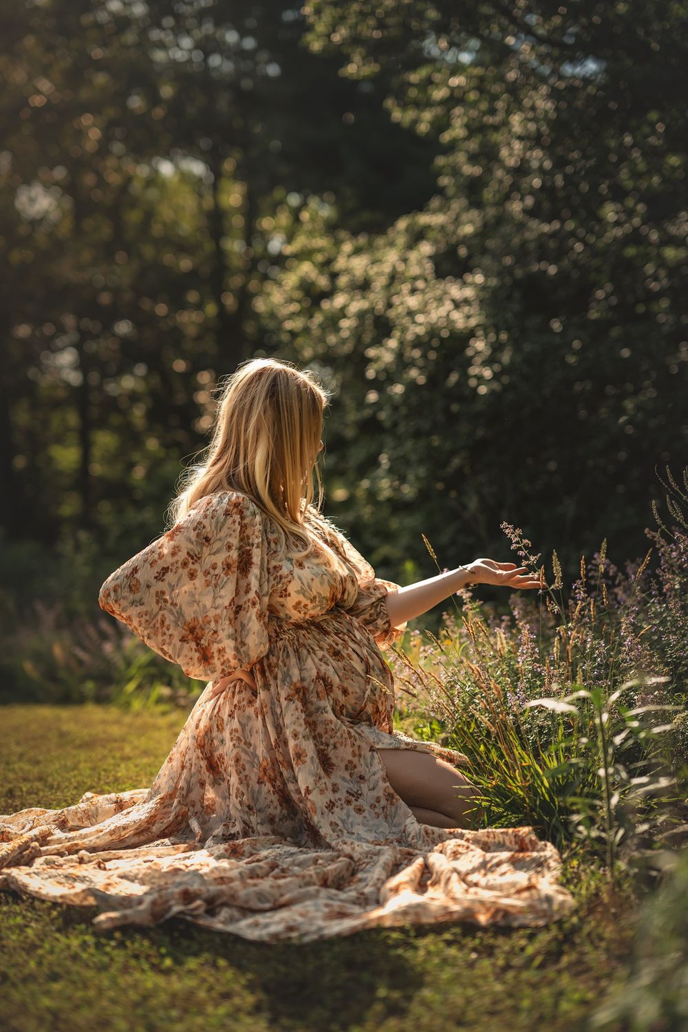 A pregnant woman in a floral dress sits in a sunlit garden reaching toward wildflowers on a peaceful summer day for maternity photos in TN.