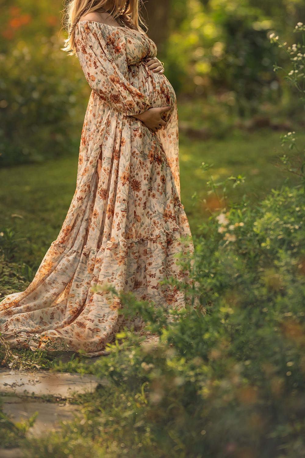 Pregnant woman in backlit floral dress flowing in the sunlit garden surrounded by green foliage creates a dreamy outdoor aesthetic.