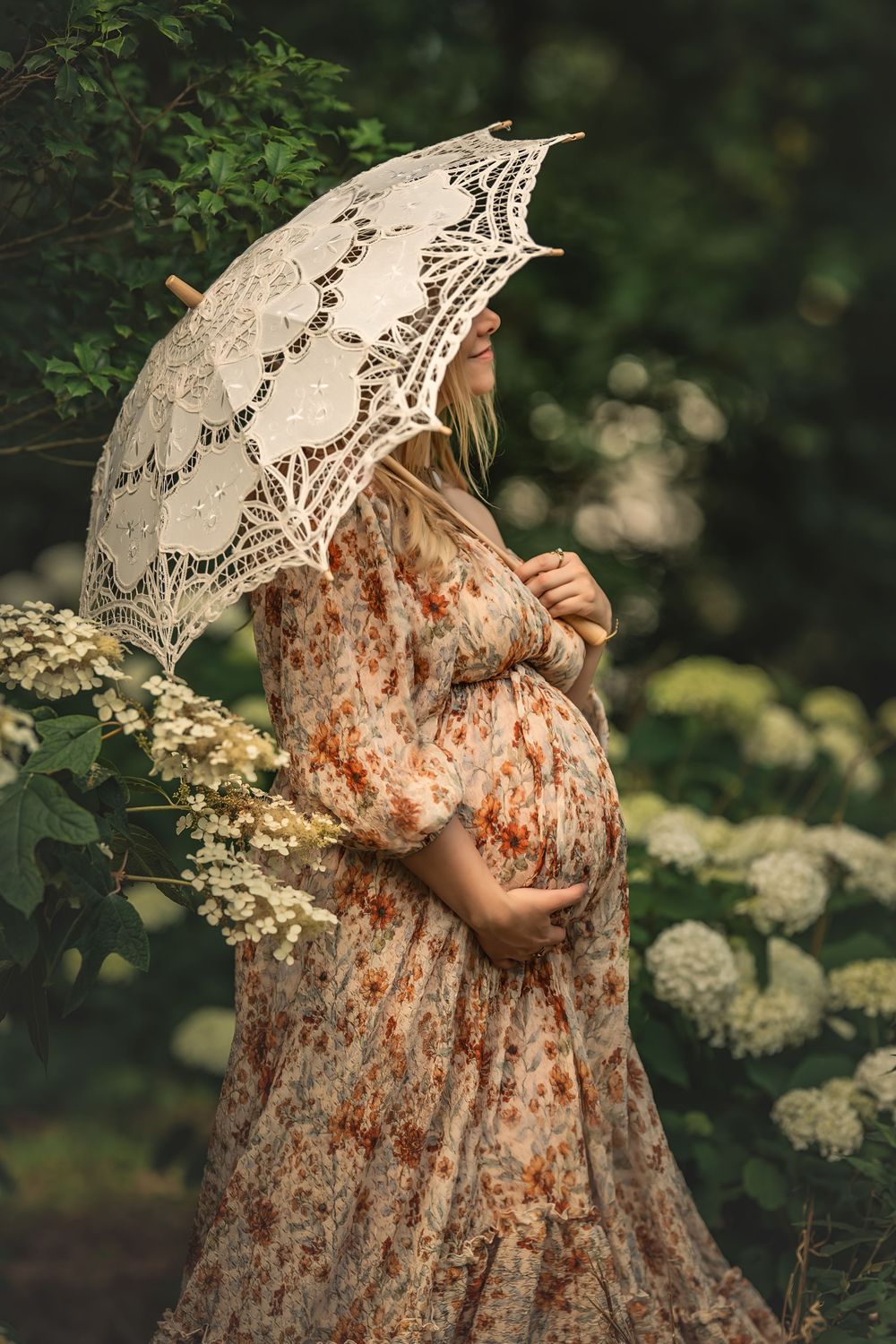 Delicate white lace parasol held over a floral dress for maternity photos in a garden with blooming hydrangeas and greenery.