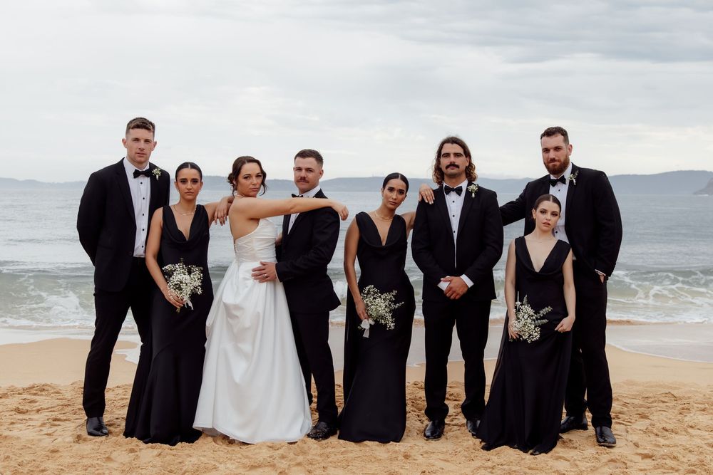 Wedding party in formal attire poses together on a beach with ocean waves and cloudy sky in the background.