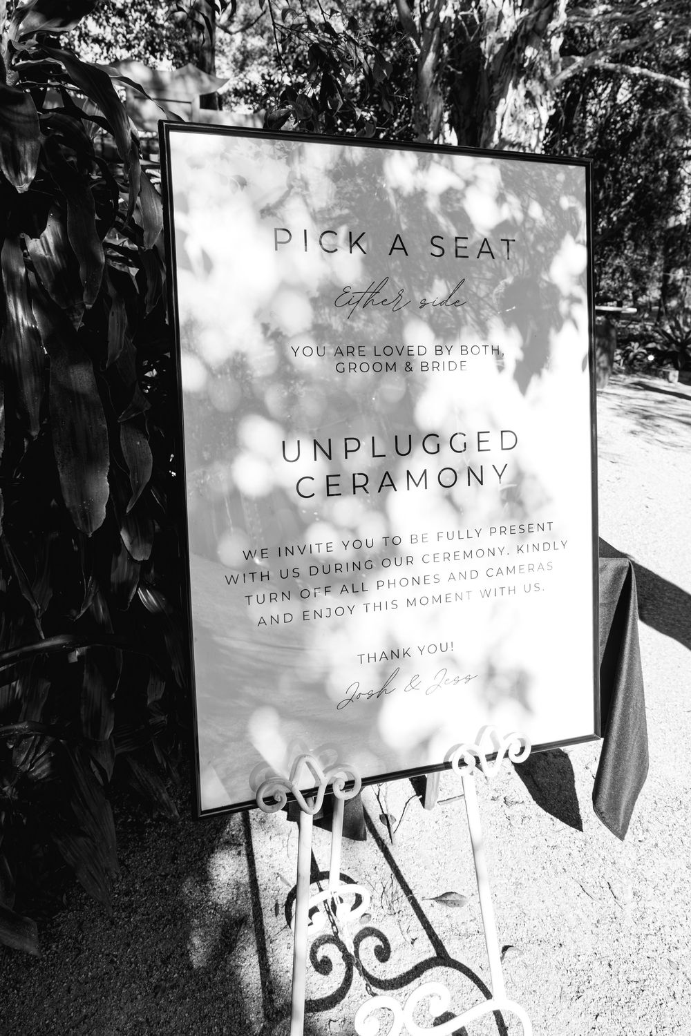 Black and white wedding ceremony signs with text displayed on white boards in an outdoor garden setting.