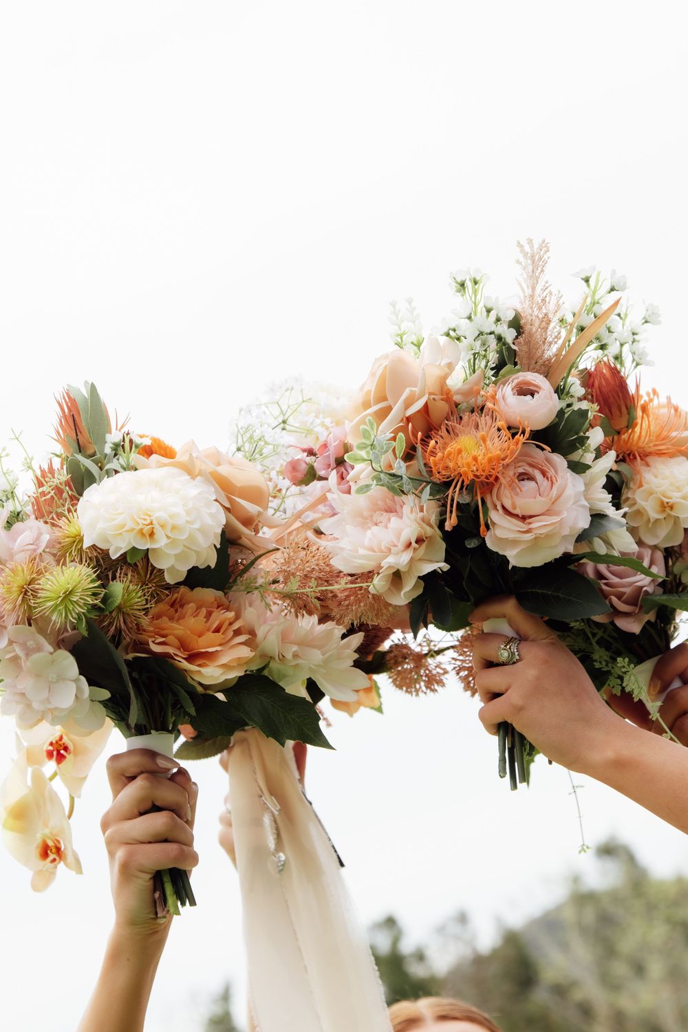 Four elegant bridal bouquets held up against a bright sky, featuring blush pink roses, white peonies and delicate greenery.