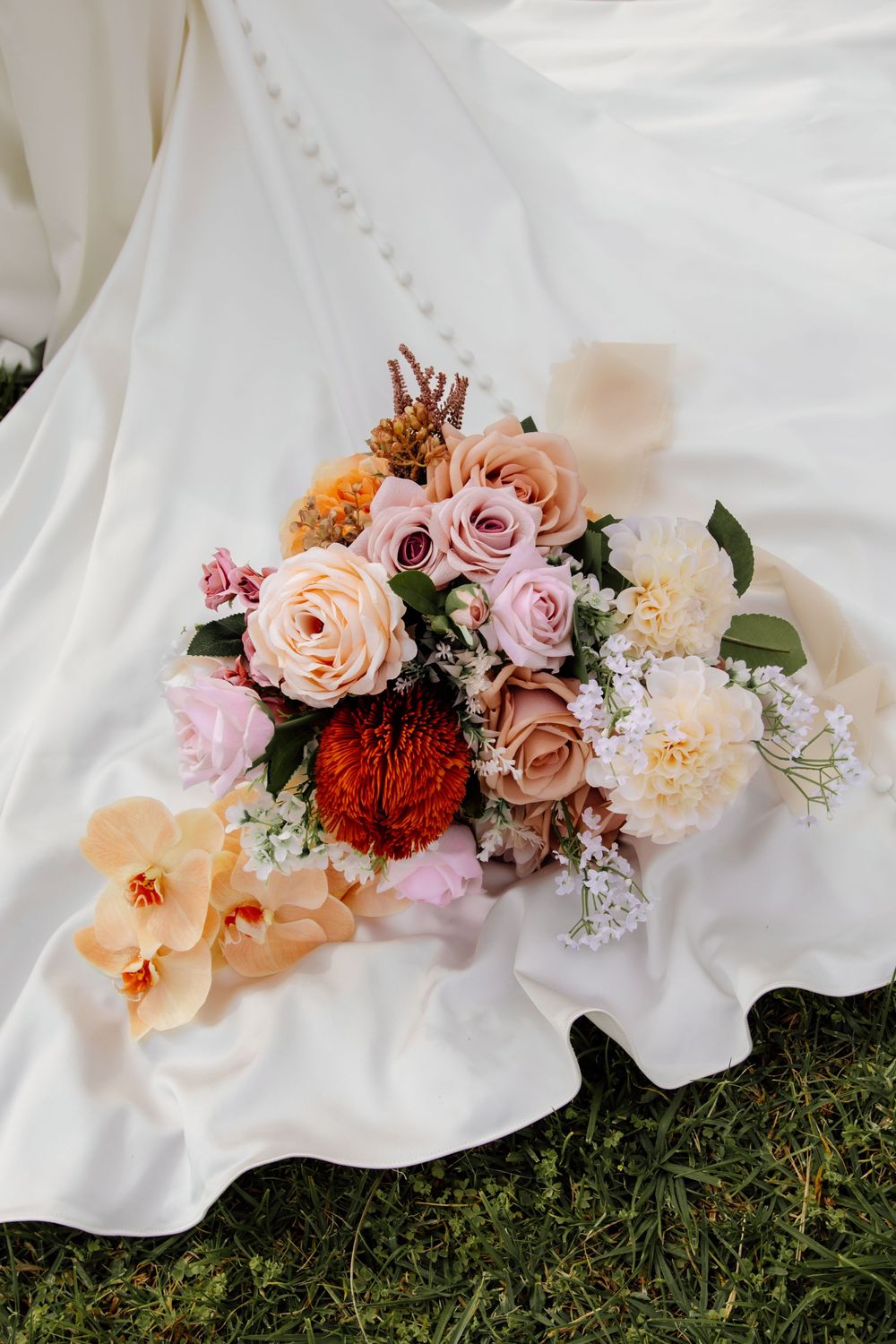Beautiful wedding bouquet with pink and peach roses, dahlias, and orchids laying on white fabric against grass.