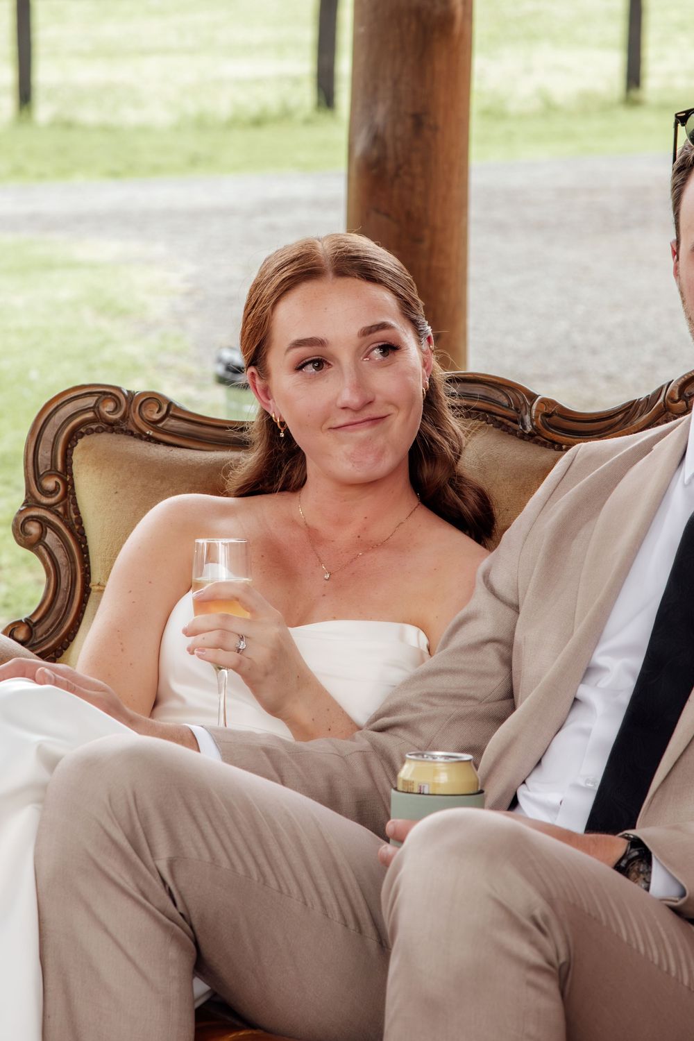 A bride in a white dress holds a champagne glass while relaxing on an ornate vintage chair at an outdoor reception.