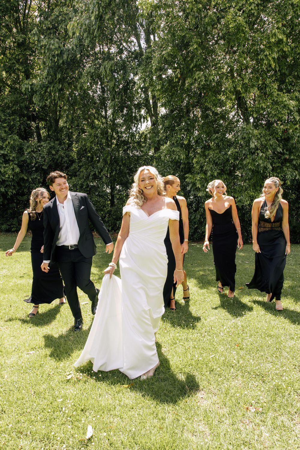 Wedding party in black and white attire poses playfully in sequence across grassy lawn with trees in background.