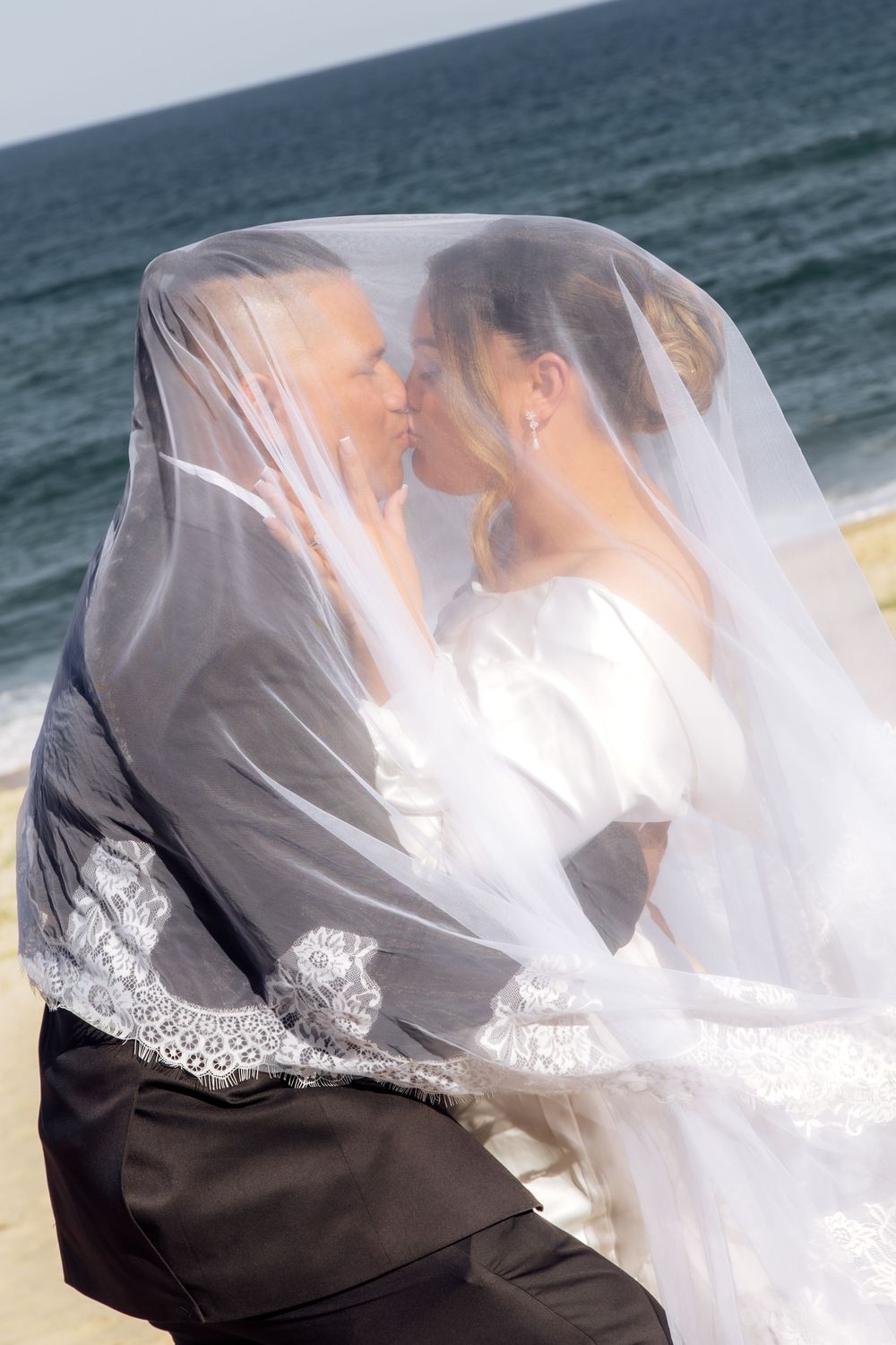 A romantic beach wedding scene with a couple embracing under a flowing white veil against an ocean backdrop.