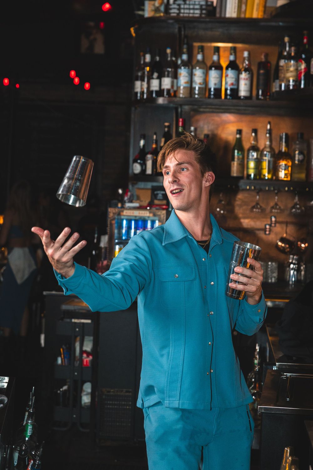 Bartender in bright blue uniform performing flair moves with cocktail shakers at a bar.