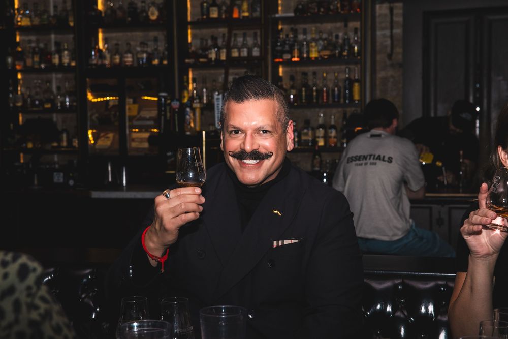 Bartender in black uniform holding up a drink at dimly lit bar.