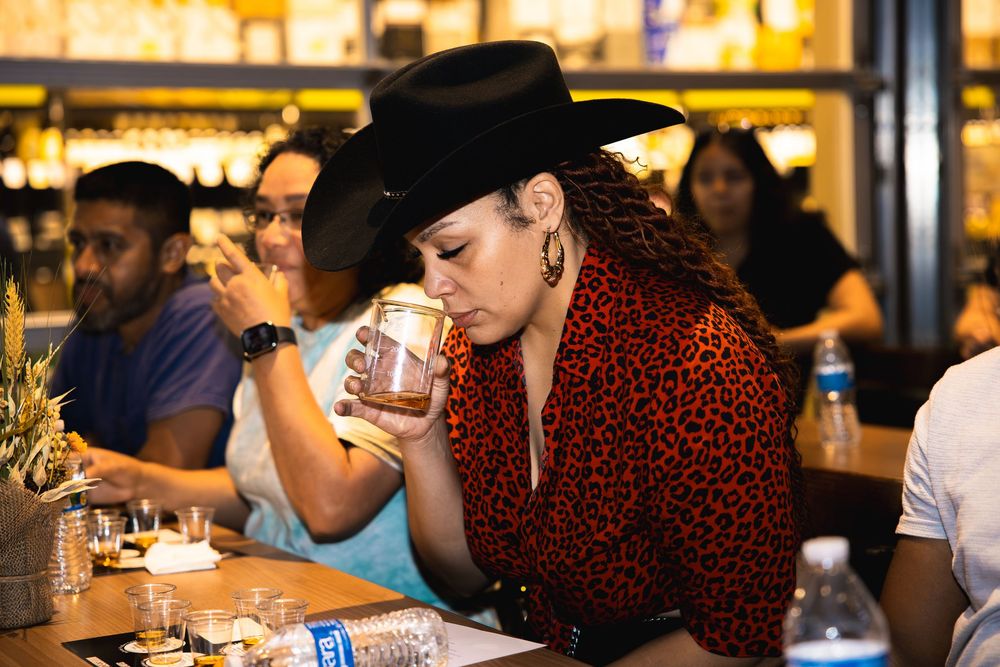 Person in black cowboy hat and leopard print shirt drinking from glass at crowded bar counter.
