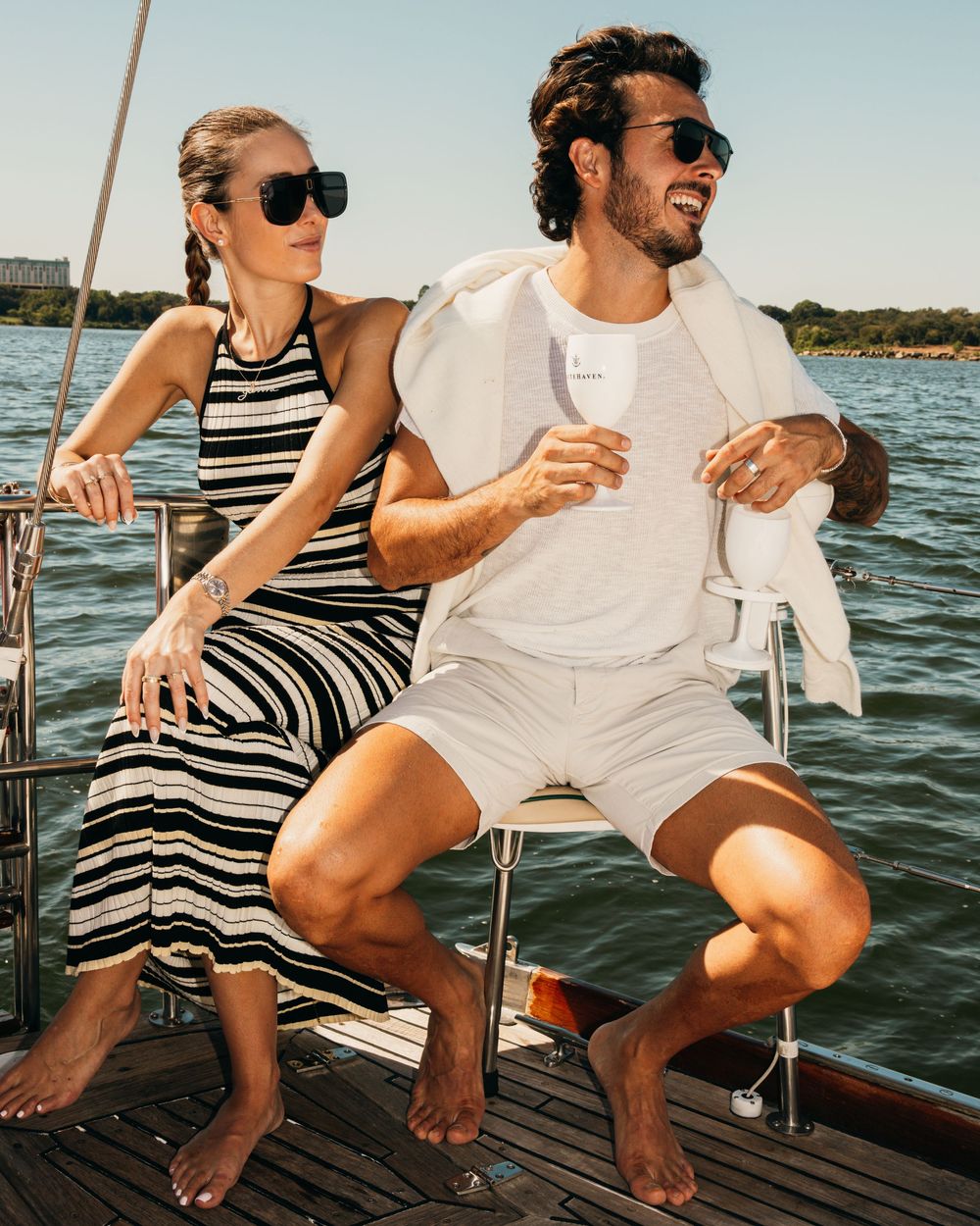 Couple enjoying drinks while relaxing on sailboat deck in summer attire.